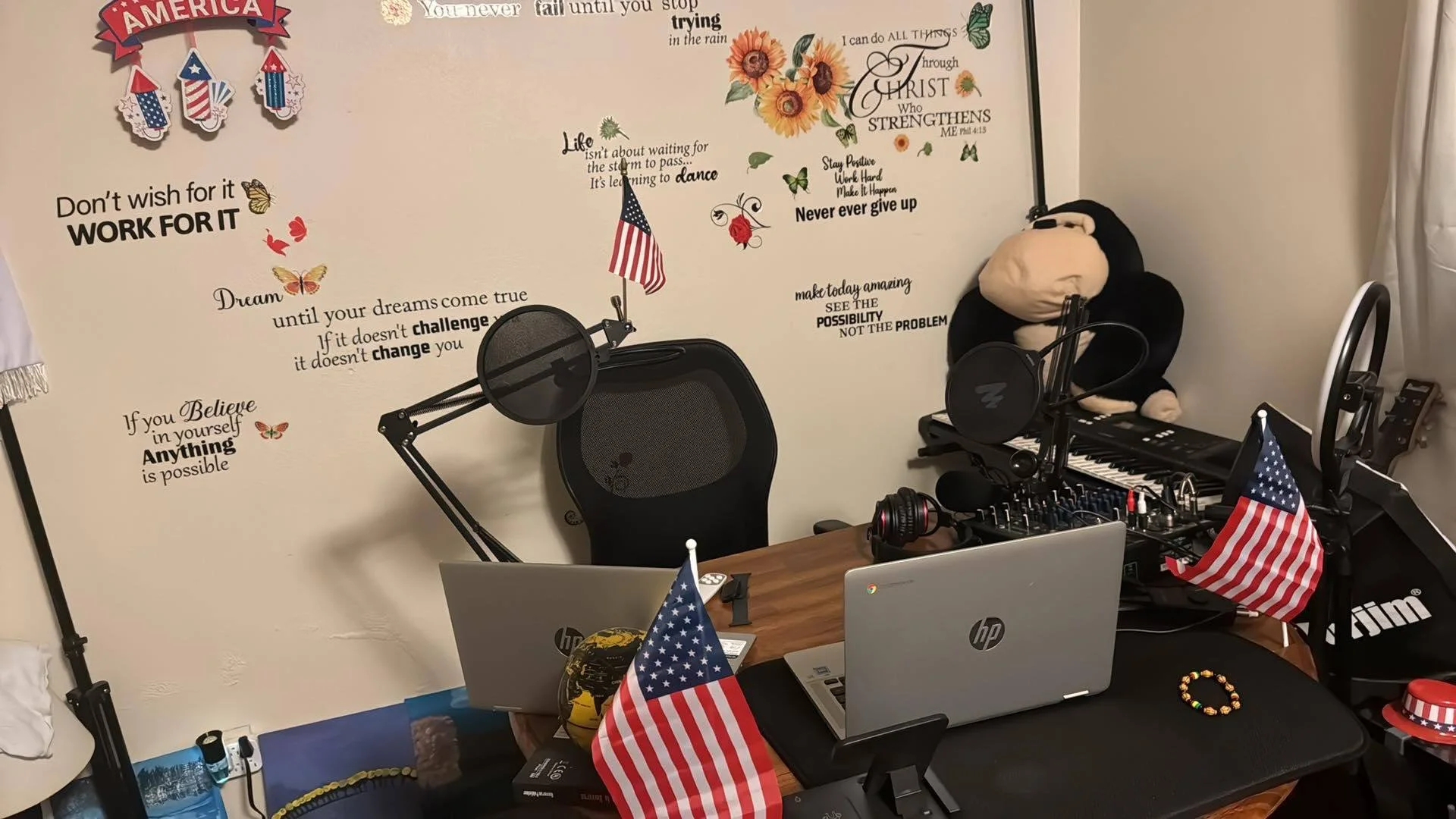 A home office decorated with American patriotic theme, featuring a wall decorated with motivational and patriotic quotes, American flags on the desks, a black keyboard, two microphones, laptops, a plush monkey toy on a stand, headphones, a microphone boom arm, and various other objects.