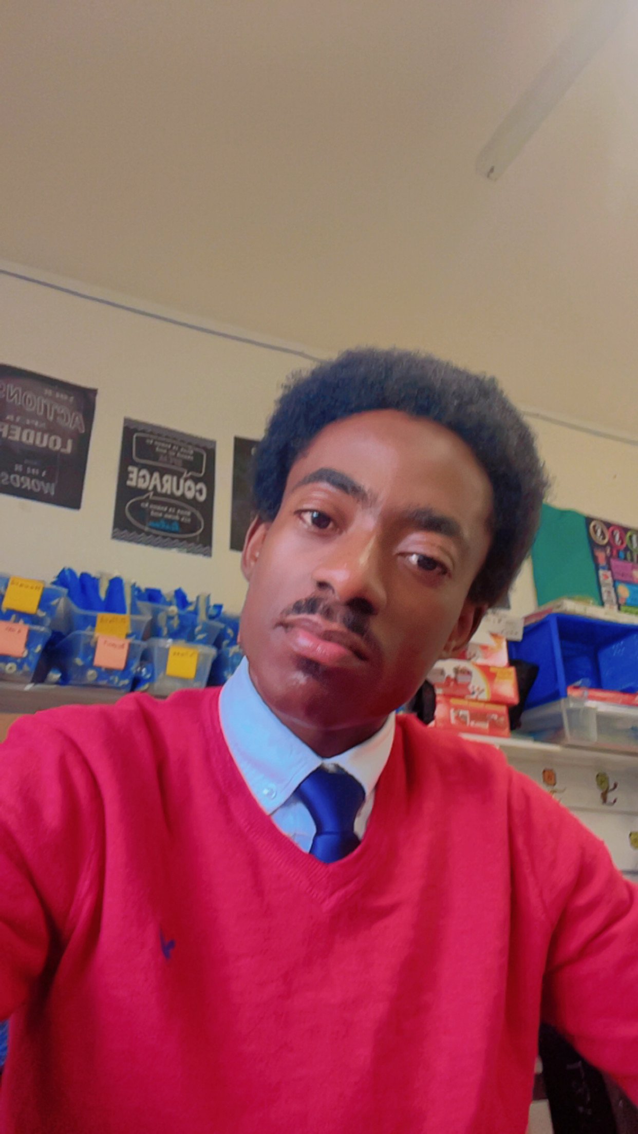 A man with short dark hair and a mustache, wearing a red sweater over a collared shirt with a blue tie, taking a selfie in a store with shelves of snacks and posters on the wall behind him.