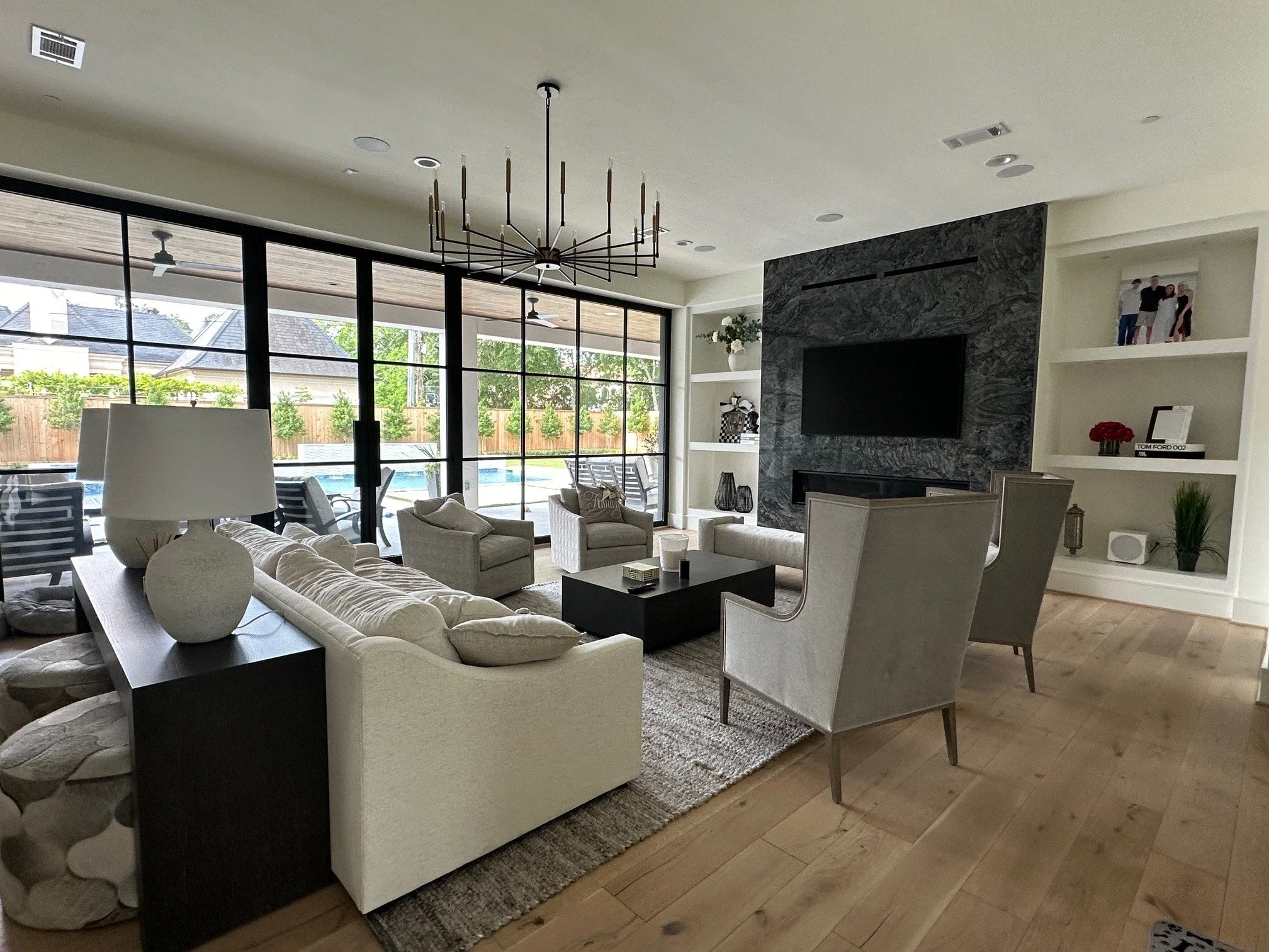 Living room with beige furniture, a black and gray marble fireplace, black built-in shelves, large glass sliding doors leading to a backyard, and a modern black chandelier on the ceiling.