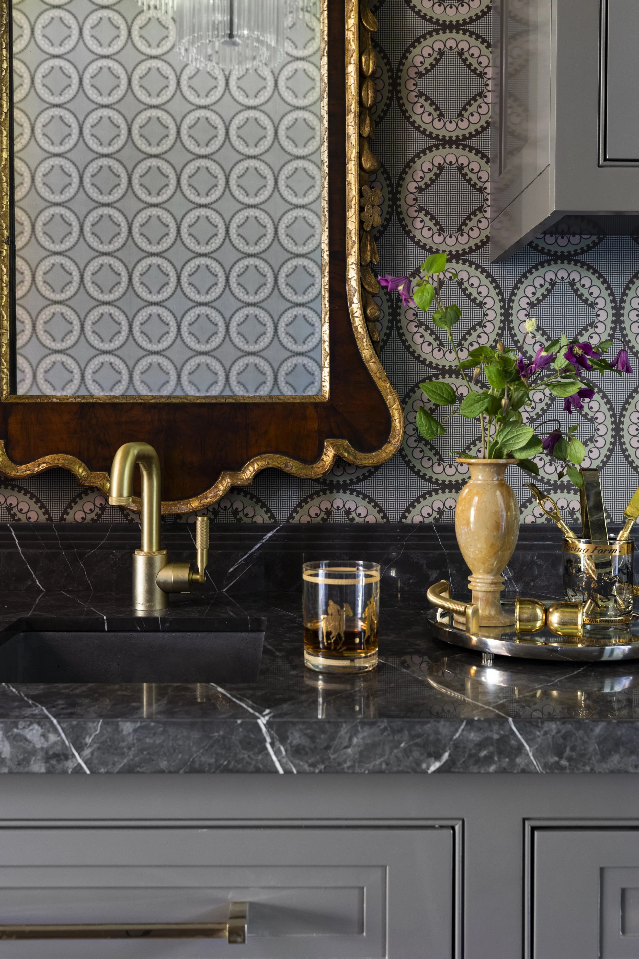 A kitchen countertop with a black marble surface featuring white veining, a gold faucet, a glass of dark beverage, a beige vase with purple flowers, and gold kitchen utensils and accessories. The background includes a patterned wallpaper and a rectangular mirror with a gold and wood frame.