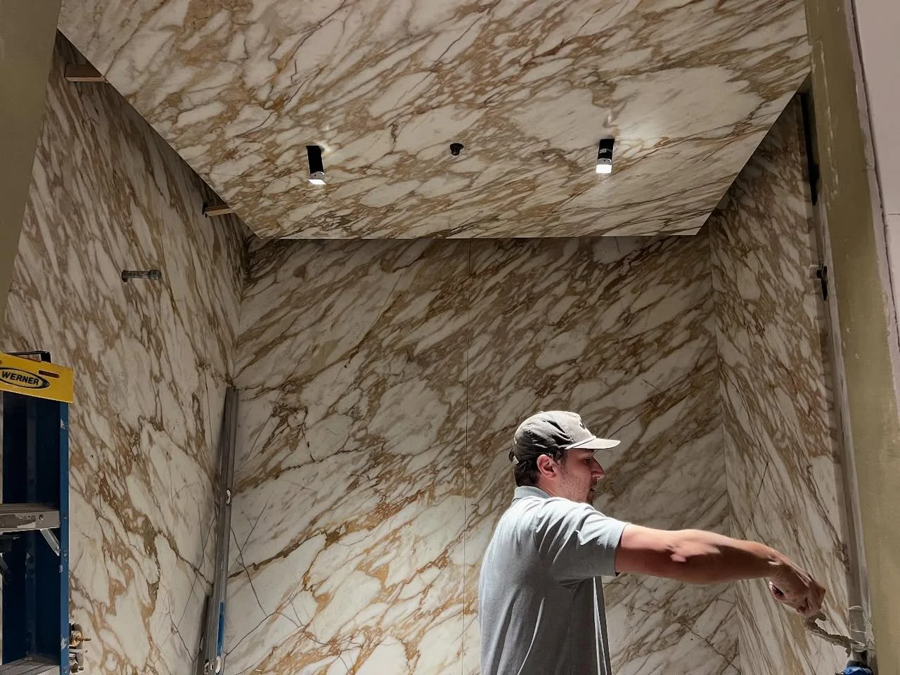 A man with a baseball cap and gray shirt working on marble wall and ceiling installation in an interior space, with a ladder on the left.