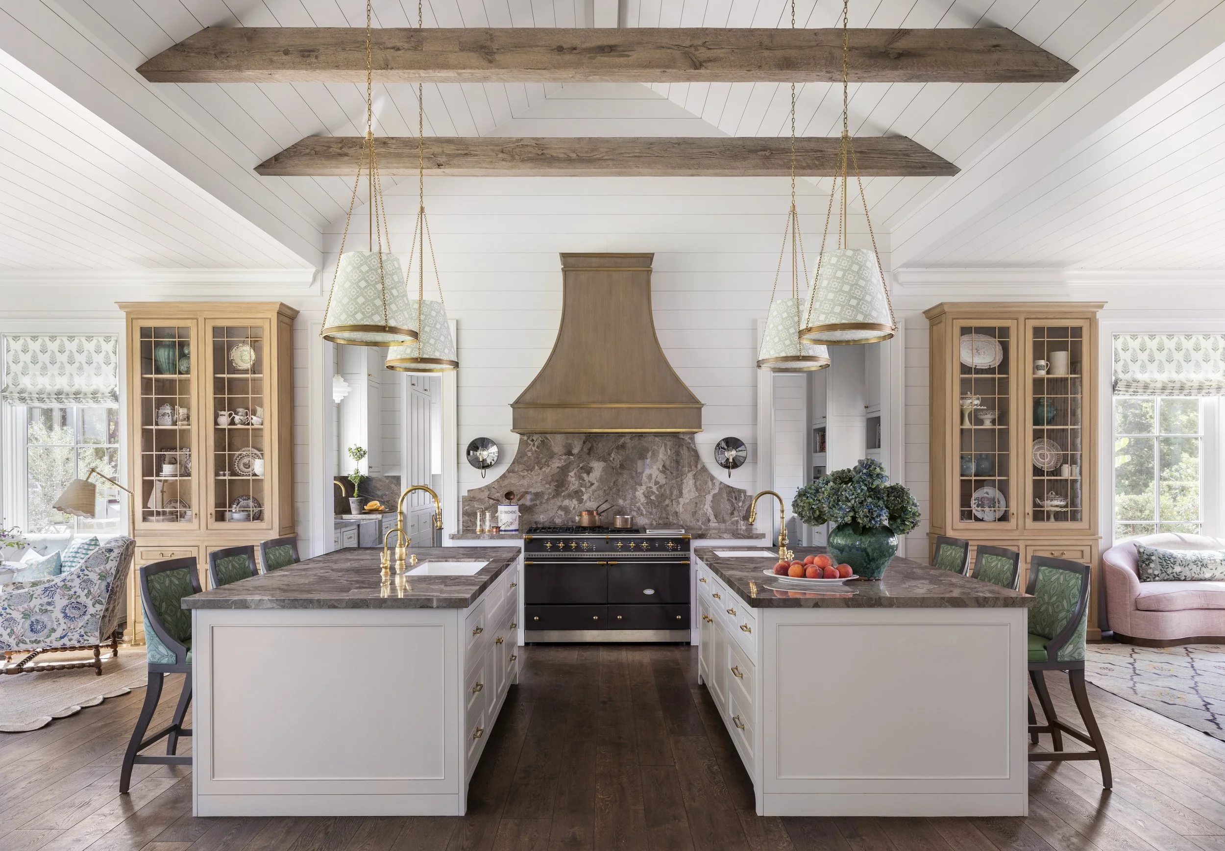 A spacious kitchen with a central island featuring a black stove and white cabinets, wooden countertops, an ornate wooden range hood, two large glass-fronted cabinets filled with dishes and decorative items, a window with patterned curtains, and modern pendant lights hanging from the ceiling.
