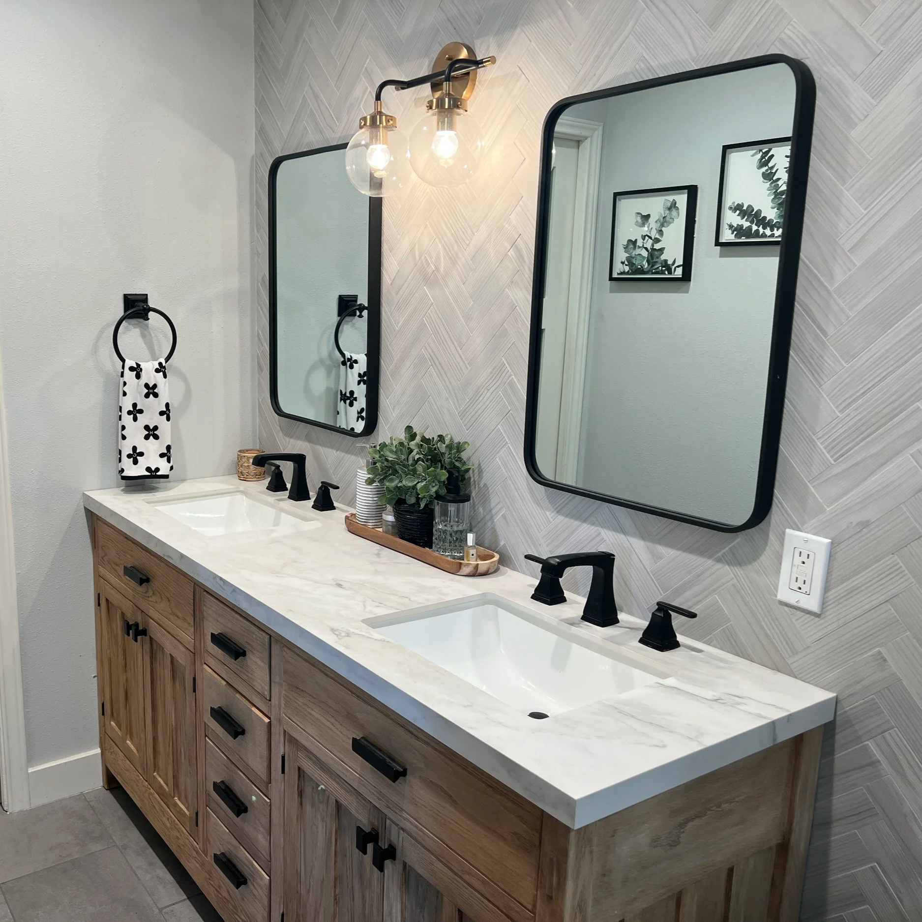 Modern bathroom vanity with a white marble countertop and black fixtures, two black framed mirrors, wall sconce lighting with two exposed bulbs, decorative plants, hand towels, and a wall outlet.