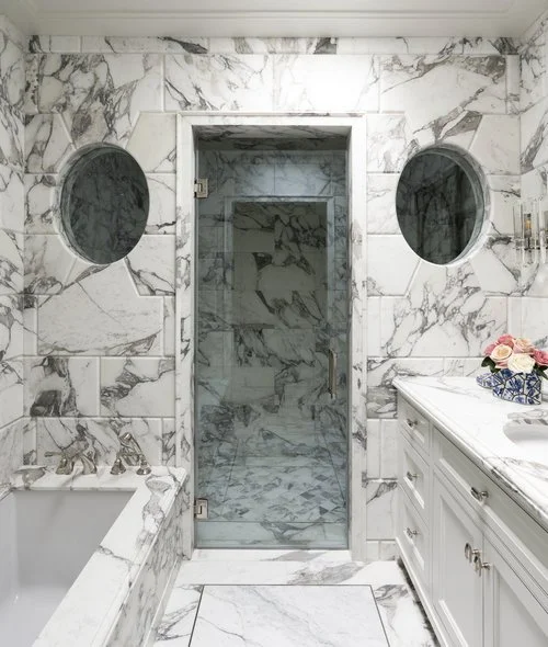 Bathroom with white marble walls, two circular mirrors, a glass shower door, a white vanity with gold handles, and a vase of pink flowers.