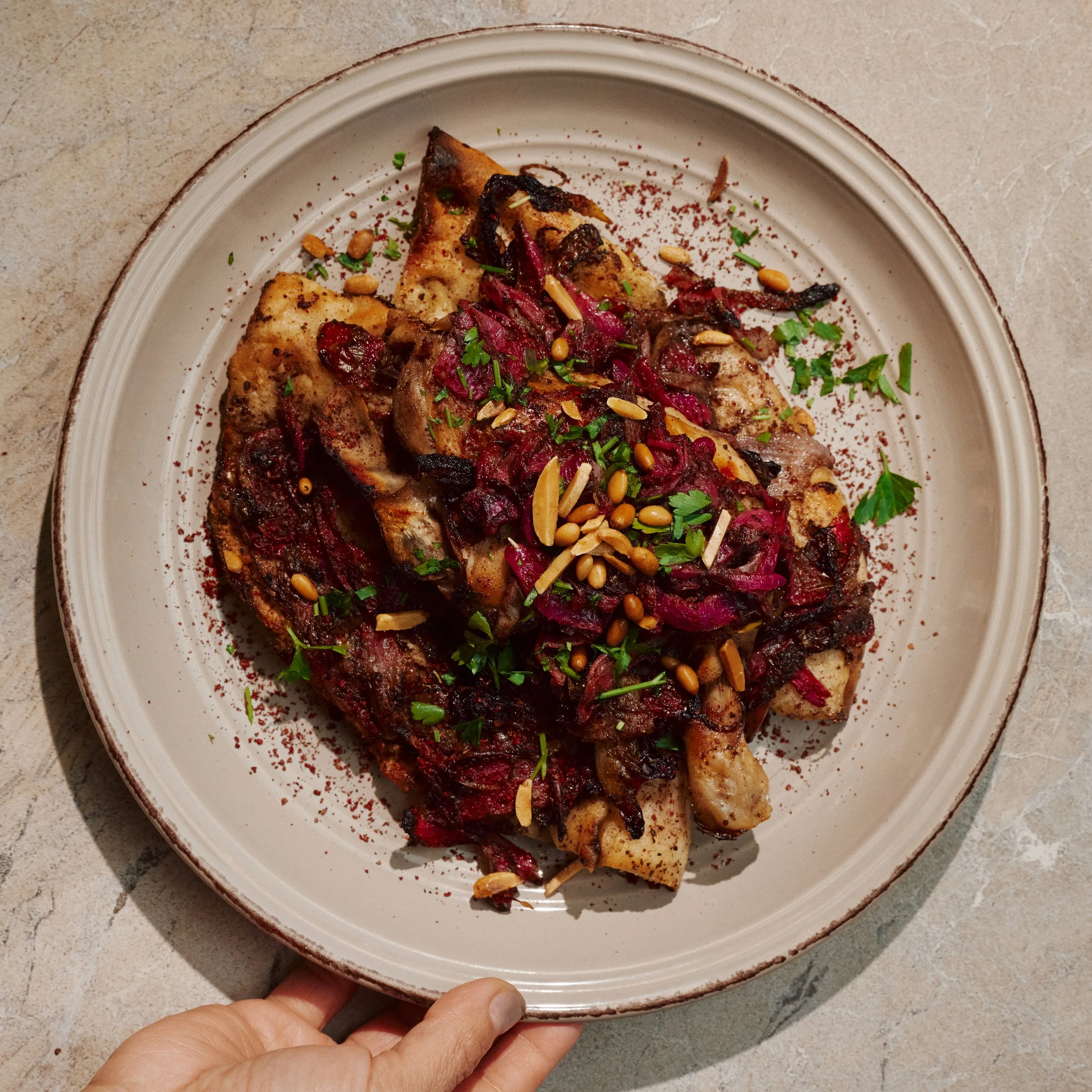 A white plate holds roasted chicken with a dark, caramelized glaze, garnished with chopped parsley, toasted pine nuts, and dried cranberries, with a reddish spice sprinkled on the plate's rim. A hand is holding the plate from the bottom, and the dish