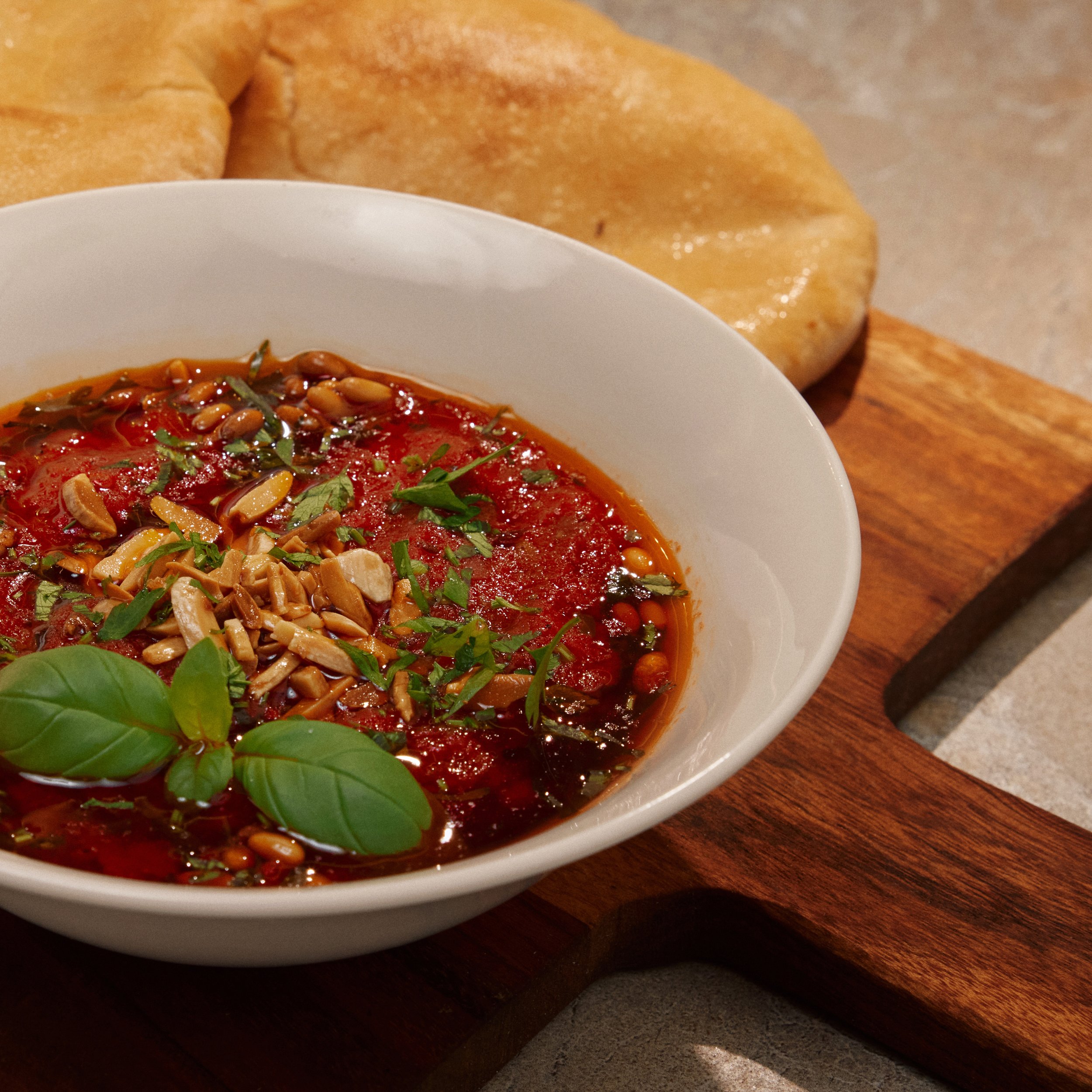 A white bowl of tomato-based soup garnished with chopped herbs and pine nuts, placed on a wooden surface with a loaf of bread in the background.