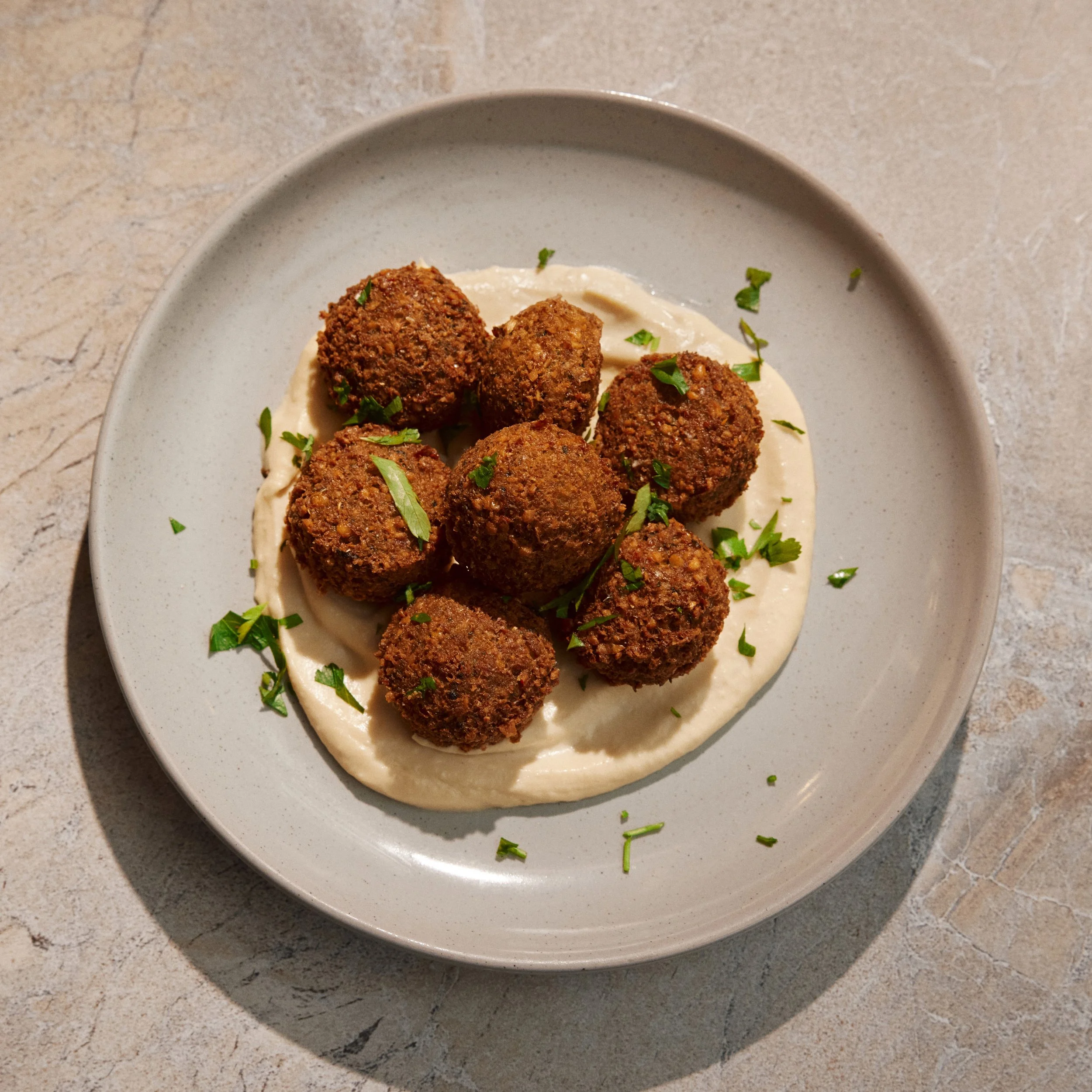 A white plate with six falafel balls garnished with chopped parsley, served on a bed of creamy sauce, on a beige textured surface.