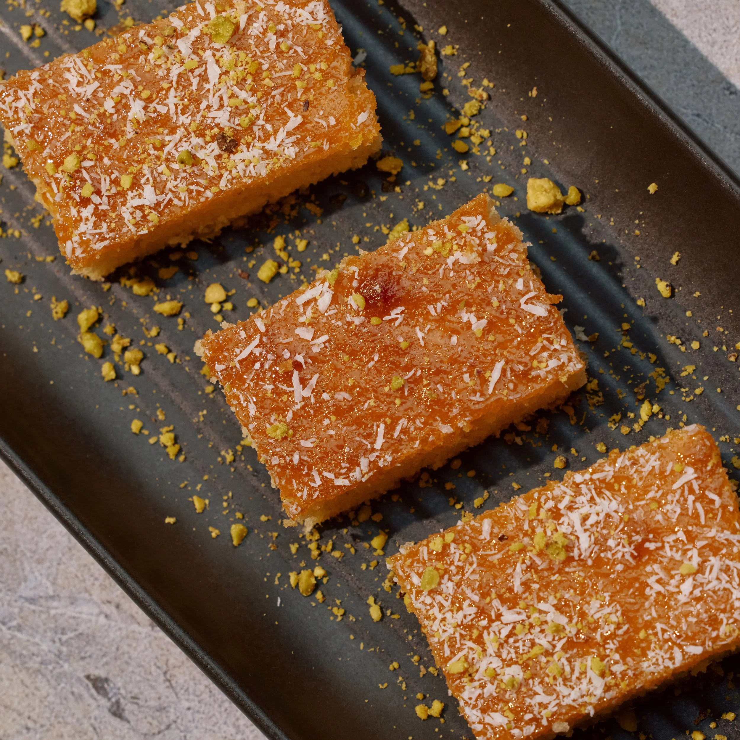 Close-up of a rectangular piece of orange dessert topped with white shredded coconut and crushed yellow pistachios, on a dark baking tray with scattered crumbs.