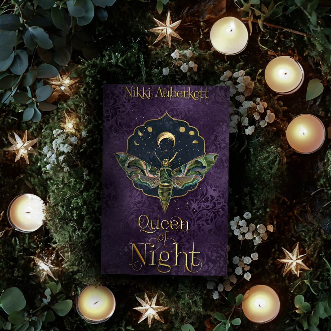 A purple book titled 'Queen of Night' by Nikki Auberkett with a moth illustration, surrounded by candles, star-shaped decorations, and foliage in a dark, cozy setting.