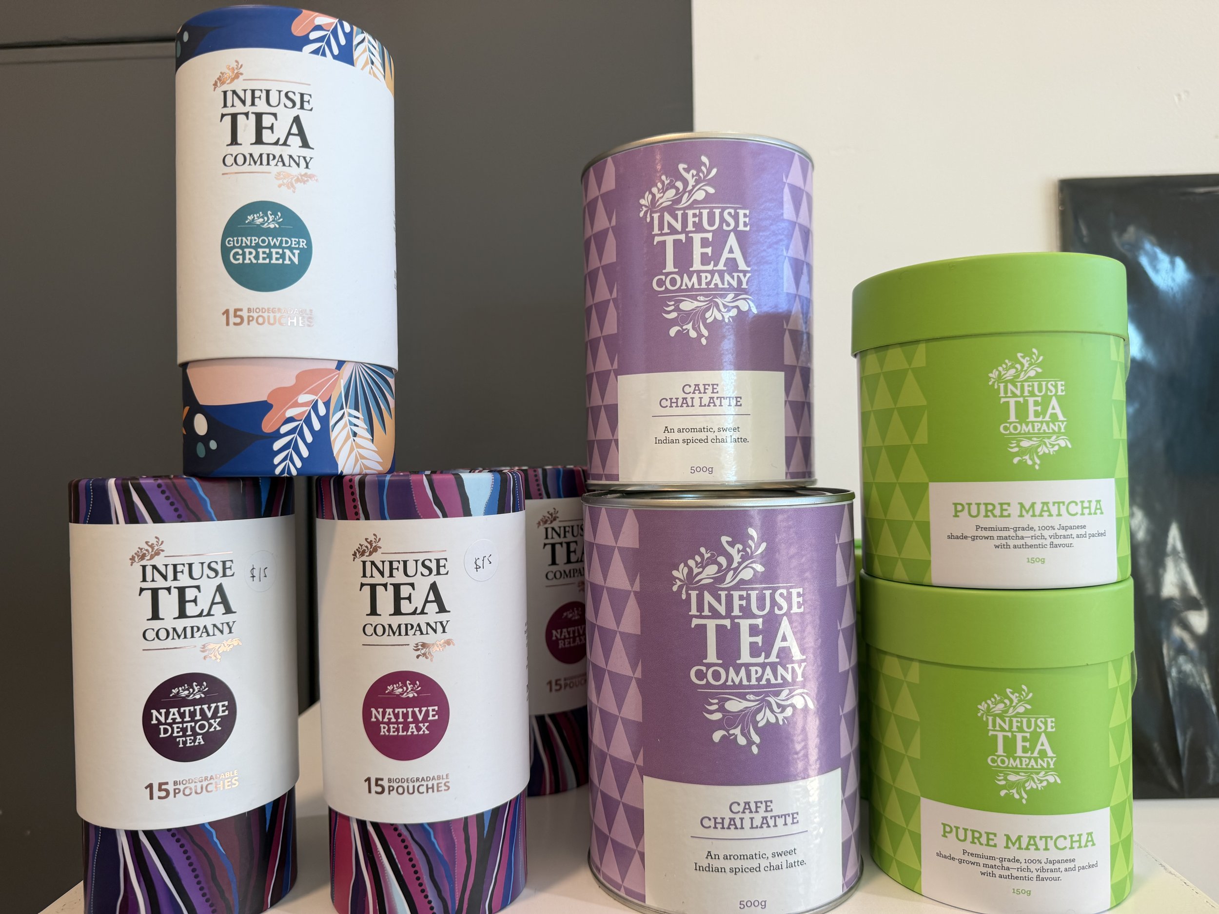Various colorful cylindrical containers of Infuse Tea Company products, including Green Gunpowder Tea, Cafe Chai Latte, and Pure Matcha, displayed on a shelf.