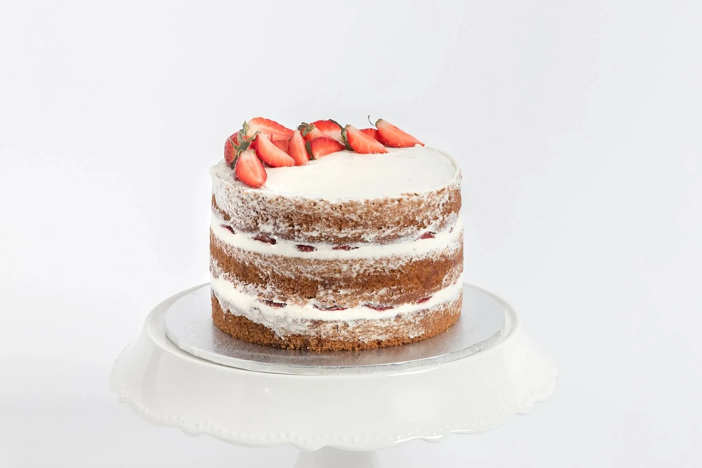 A three-tiered naked cake with strawberries on top and buttercream filling, displayed on a white cake stand against a white background. Buttercream cake. Strawberry cake. vegan cake. cakes in exmouth. exmouth cake