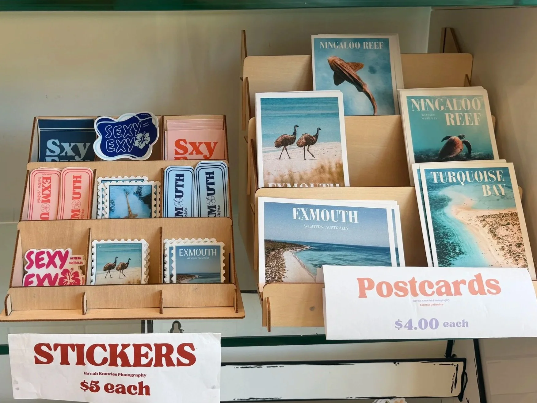 Rah Rah Collective Stickers and Postcards. Display of postcards and stickers featuring beach scenes and Australian wildlife, with signs indicating prices of $4.00 for postcards and $5.00 for stickers. Ningaloo gifts