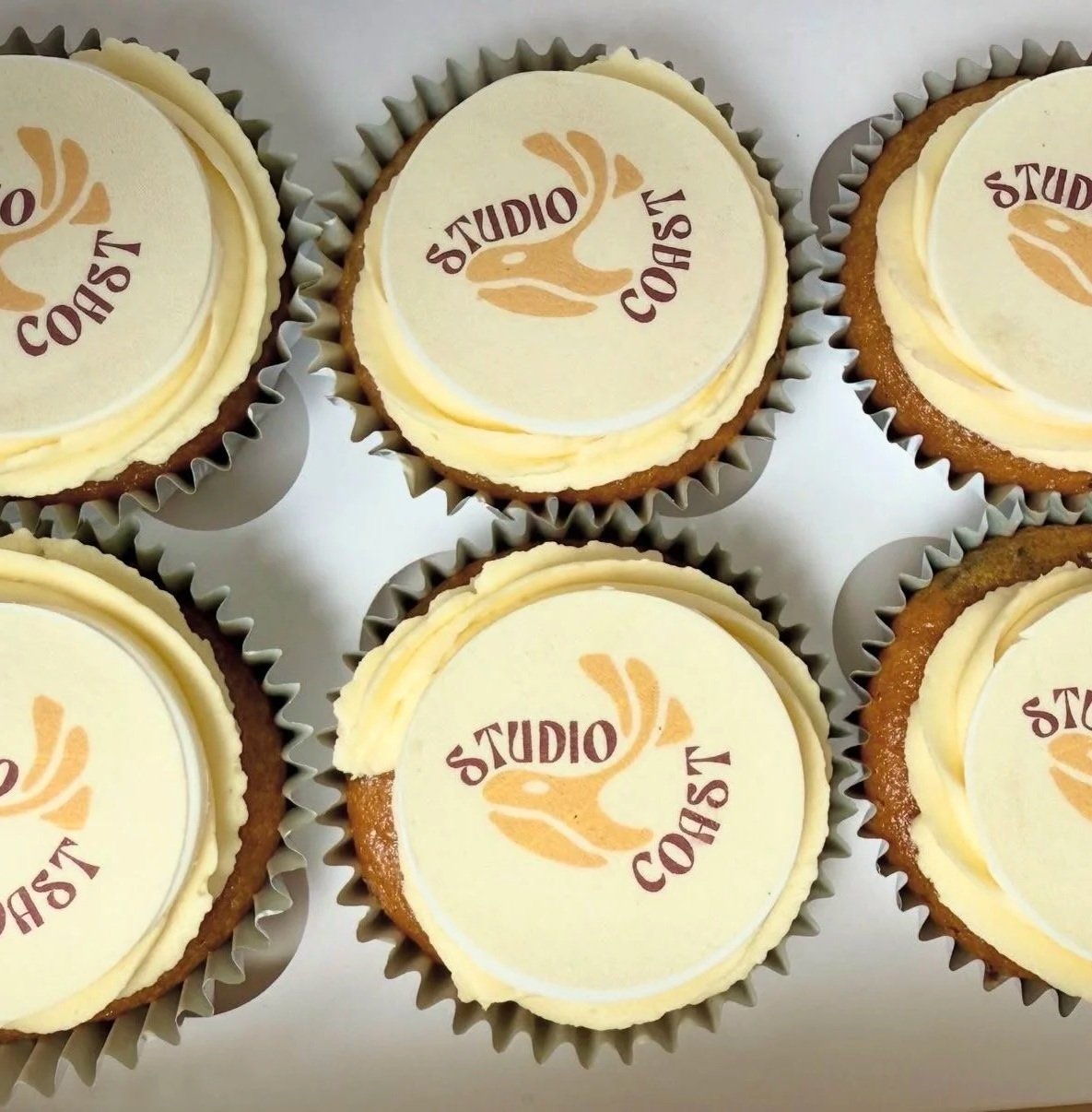 Assorted cupcakes with a logo on top that reads 'Studio Coast' in brown and orange colors. Buttercream cupcake. vegan cupcake. cupcakes in exmouth. exmouth cupcakes. Edible print cupcake toppers