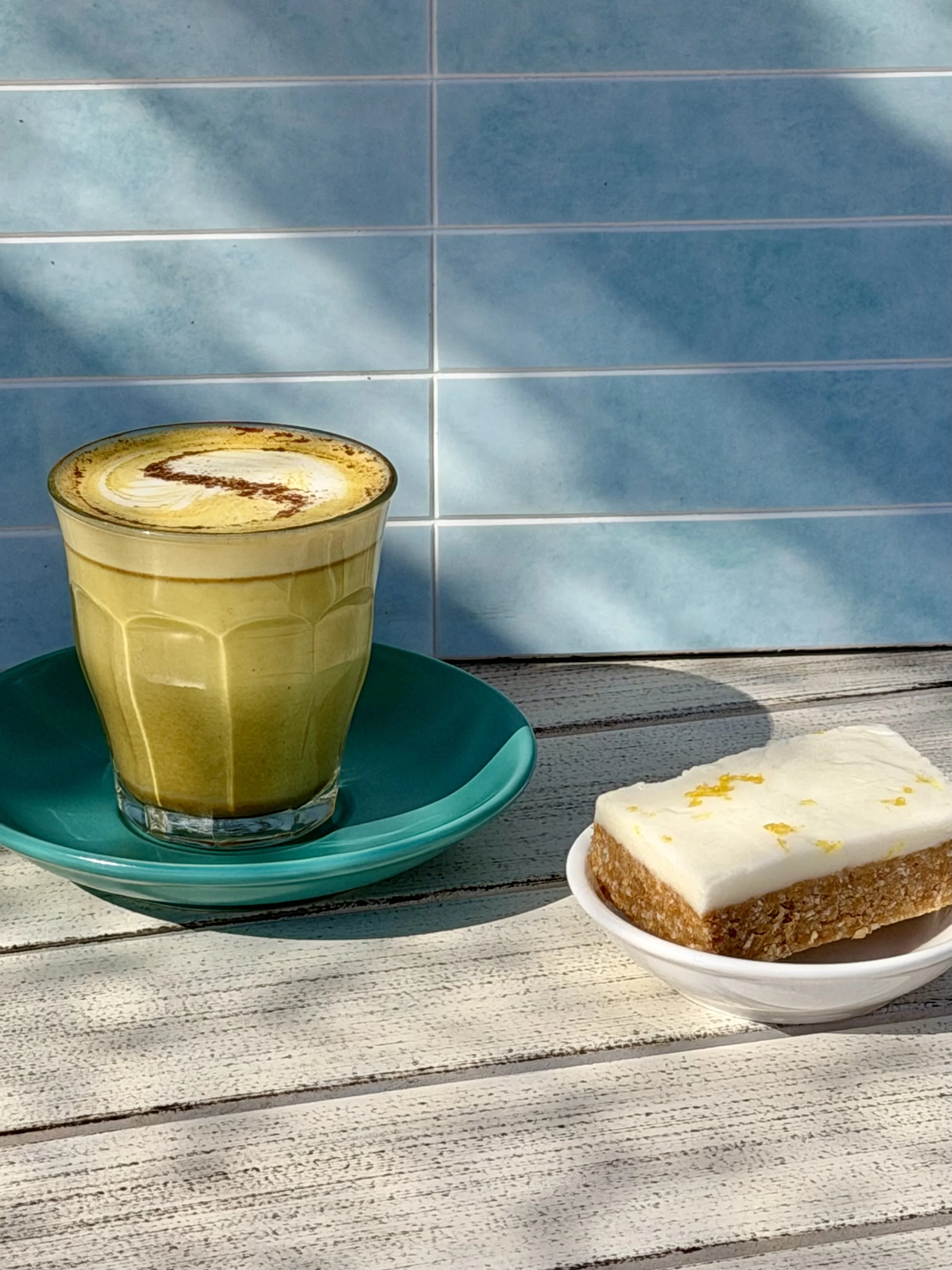 A turmeric  latte in a transparent glass on a green plate, beside a lemon slice, all placed on a rustic wooden table outdoors, with blue tile wall in the background.