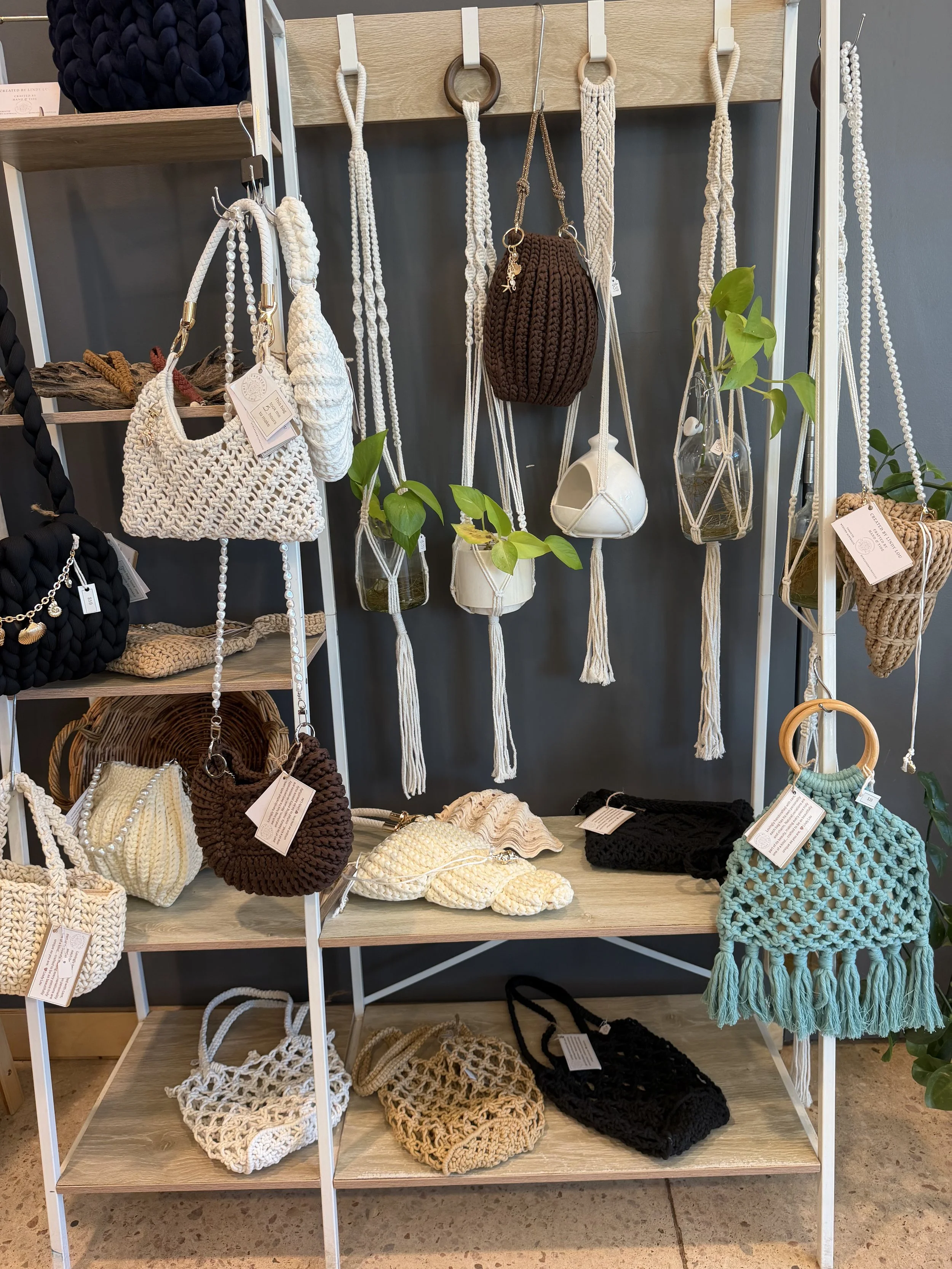 Display of handmade crocheted bags, baskets, and accessories on a multi-tiered shelf and hanging rod in a store. The items are in neutral colors like white, cream, brown, and black, with some greenery as decoration. Created by Lindy Lou