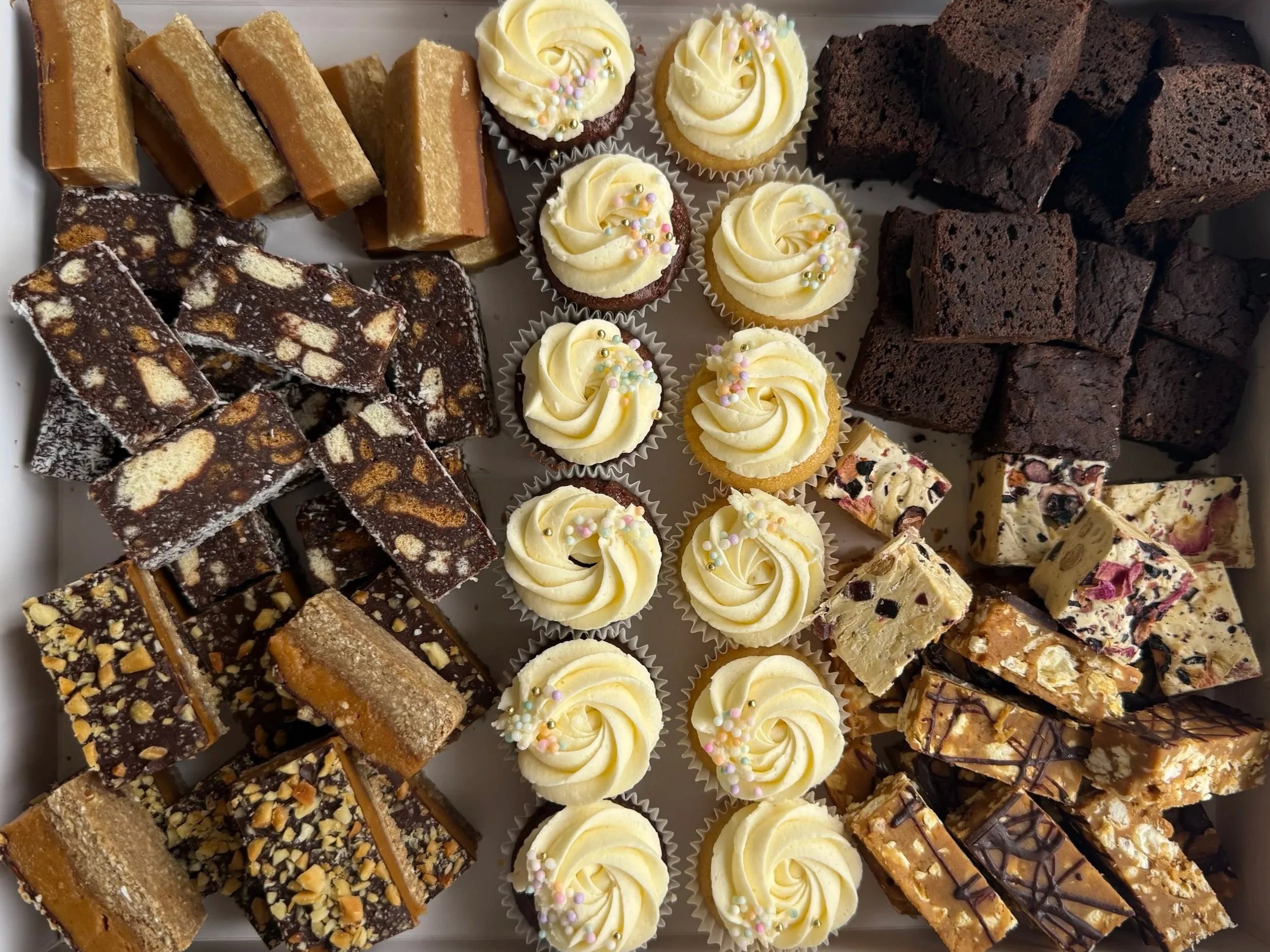 A tray of assorted desserts including chocolate-covered bars with nuts, caramel fudge, cupcakes with white frosting and sprinkles, and chocolate brownies.  catering. dessert platter. catering boxes exmouth