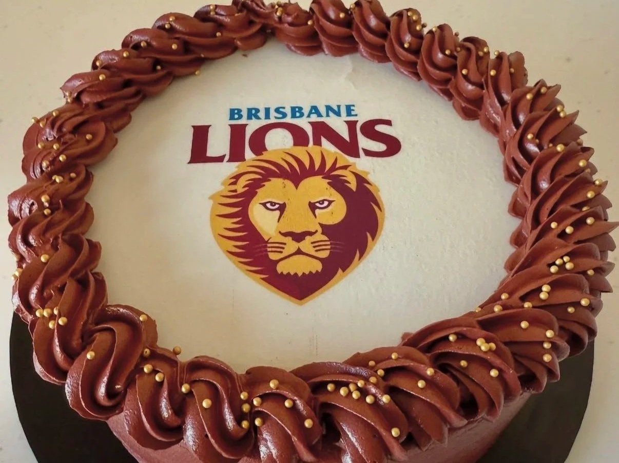 Chocolate cake with a white top featuring the Brisbane Lions team logo and name, surrounded by a border of chocolate frosting swirls. Buttercream cake. edibile print cake. vegan cake. cakes in exmouth. exmouth cake