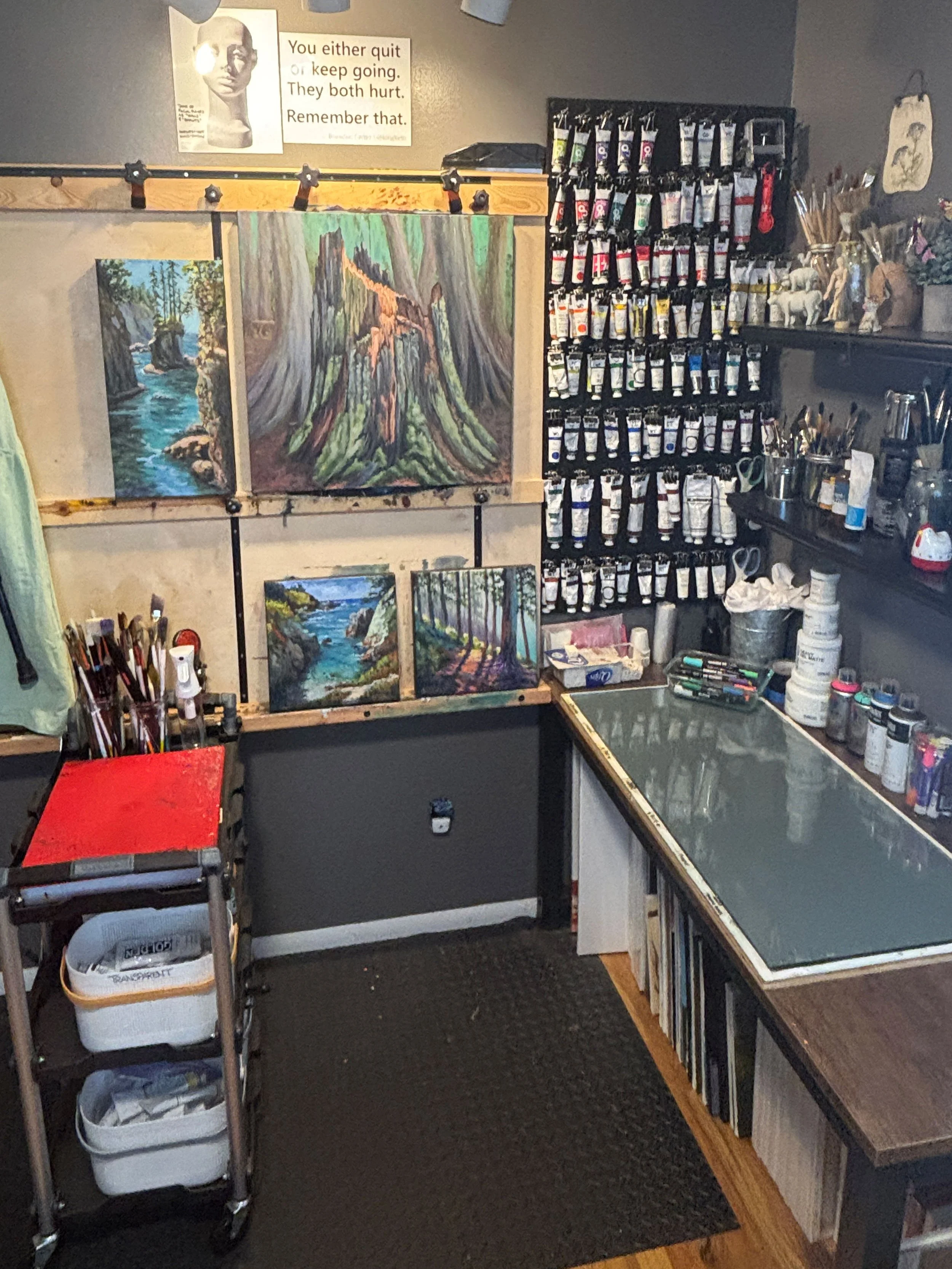 Artist’s studio with landscape paintings, paintbrushes, art supplies, and shelves of acrylic paint tubes.