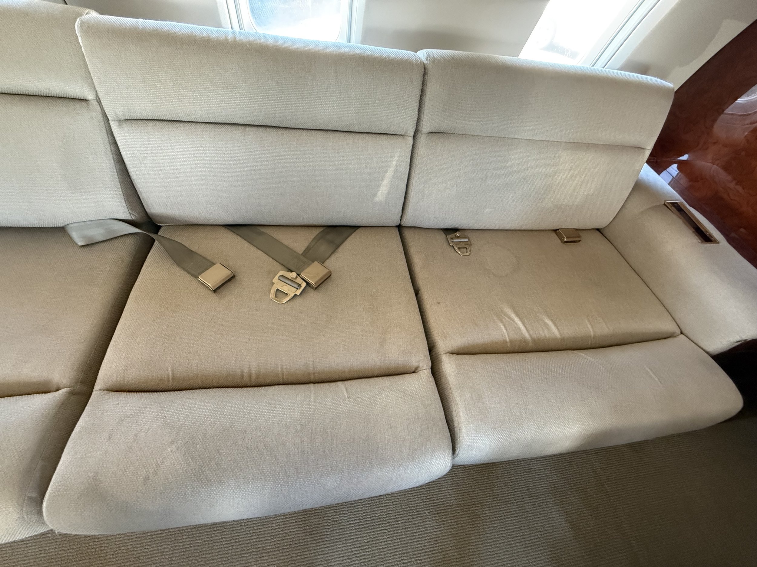 Beige fabric sofa seating inside a private jet cabin, showing visible upholstery stains, seat cushions, and safety belts before professional aircraft interior detailing.
