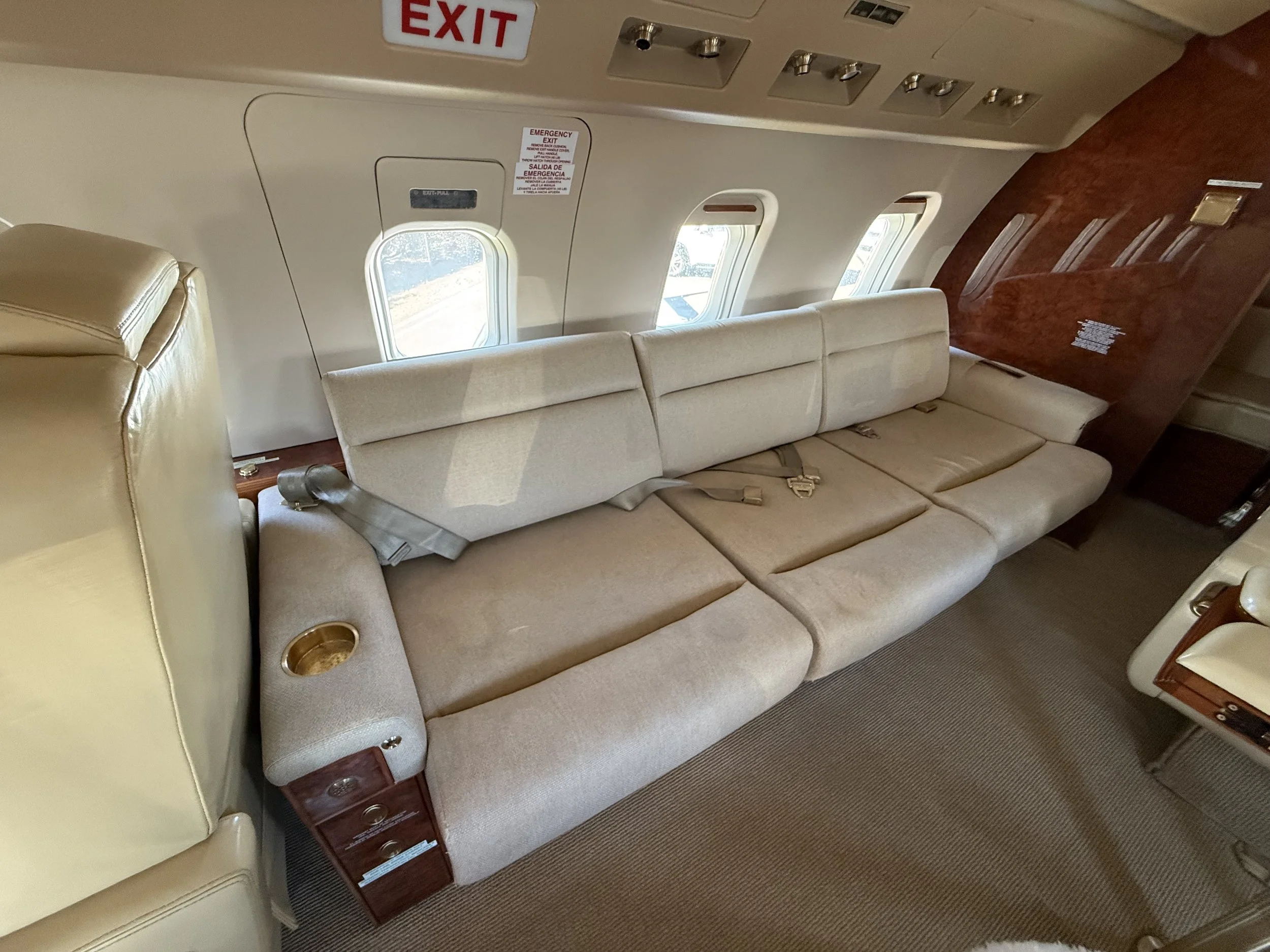 Three-seat upholstered sofa inside a Challenger 604 private jet cabin at McClellan-Palomar Airport, featuring beige seating, seatbelts, wood trim, and carpeted flooring.