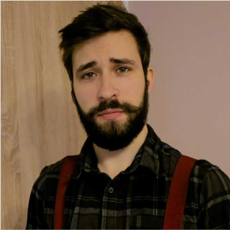 A man with dark hair and a beard, wearing a flannel shirt with suspenders, looking into the camera with a confident expression. Background features a light-colored wall and wooden paneling.