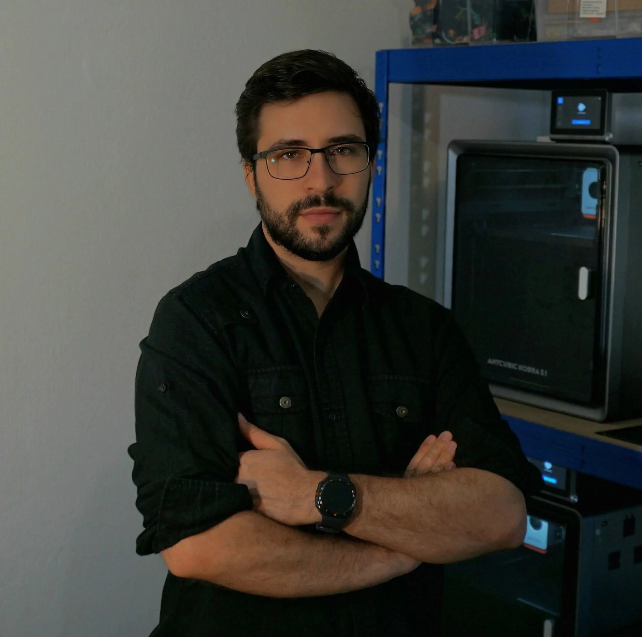A man with dark hair, beard, and glasses wearing a black shirt, standing with his arms crossed in front of a vending machine in an indoor setting.