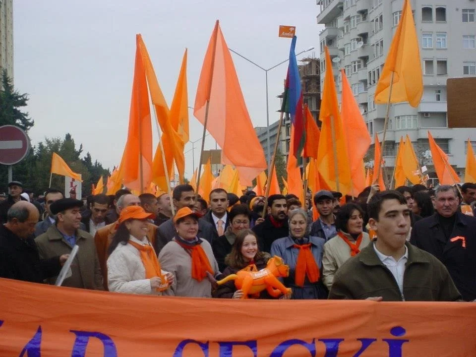 A crowd of people participating in a march or rally, holding orange flags and banners, many wearing orange scarves or hats. The event appears to be in an urban area.