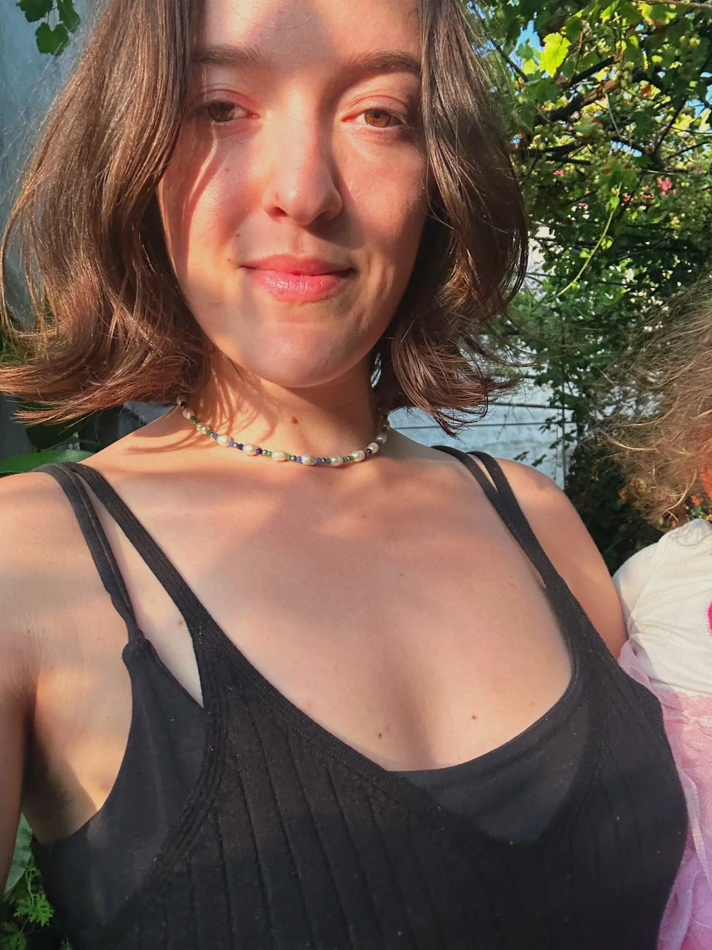 A woman with shoulder-length brown hair and light skin, smiling subtly, wearing a black top with spaghetti straps and a colorful beaded necklace, standing outdoors with green foliage and blue sky in the background.