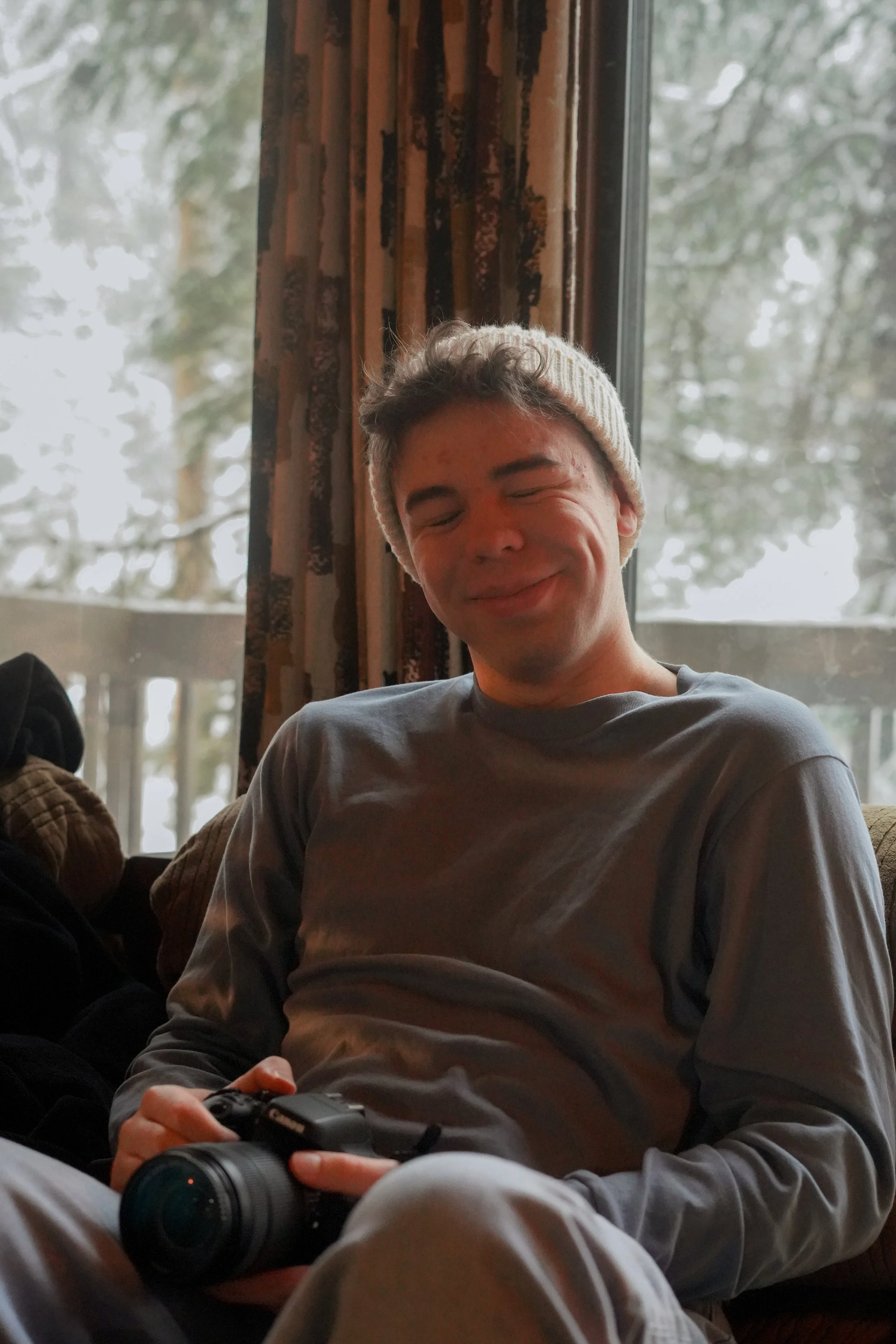 A young man with curly hair wearing a knit white beanie, smiling with eyes closed, sitting indoors near a window with trees outside. He is holding a camera in his hands and wearing a gray long-sleeve shirt.
