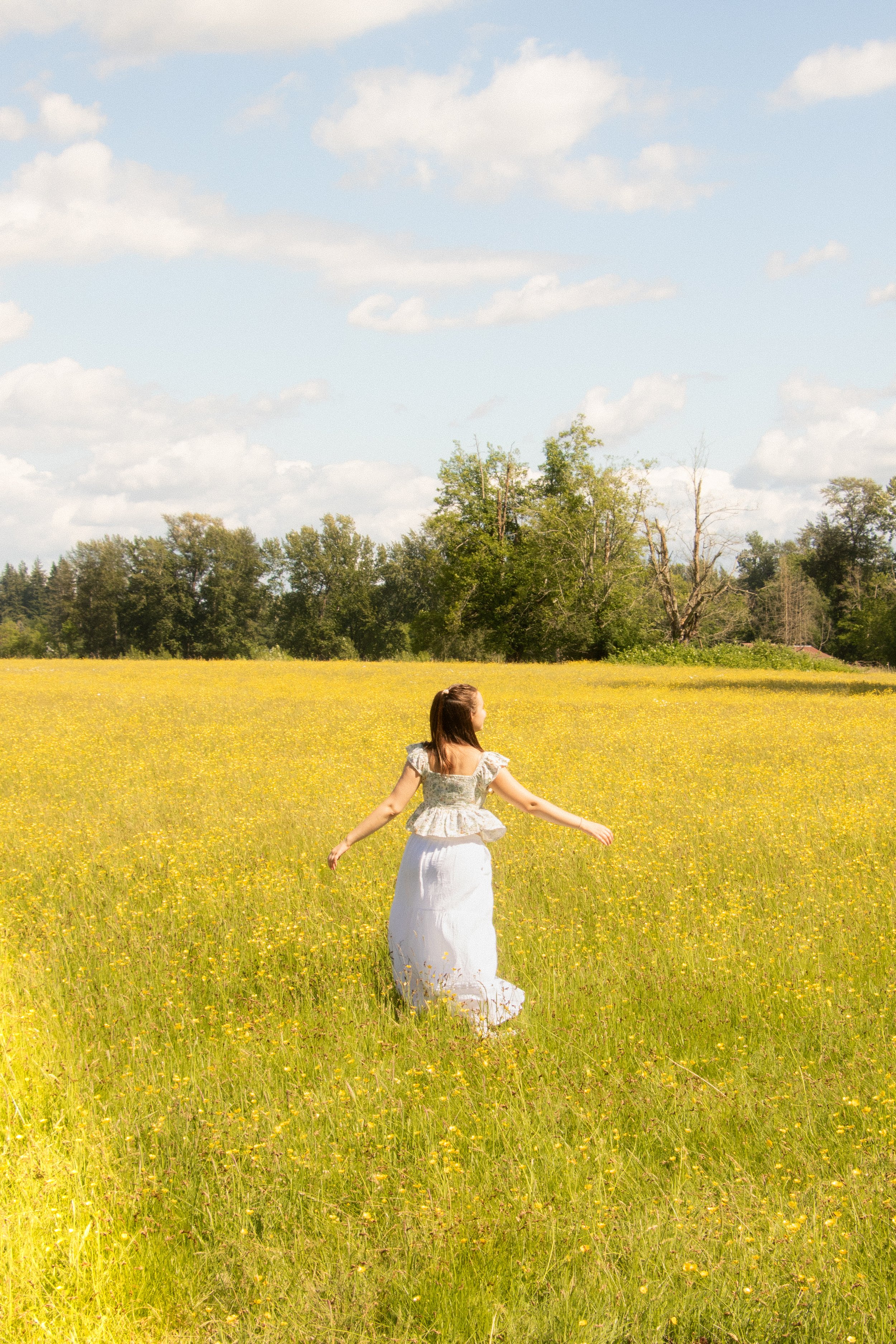 A woman in a white dress walking through a yellow flower field on a sunny day with a blue sky and scattered clouds.