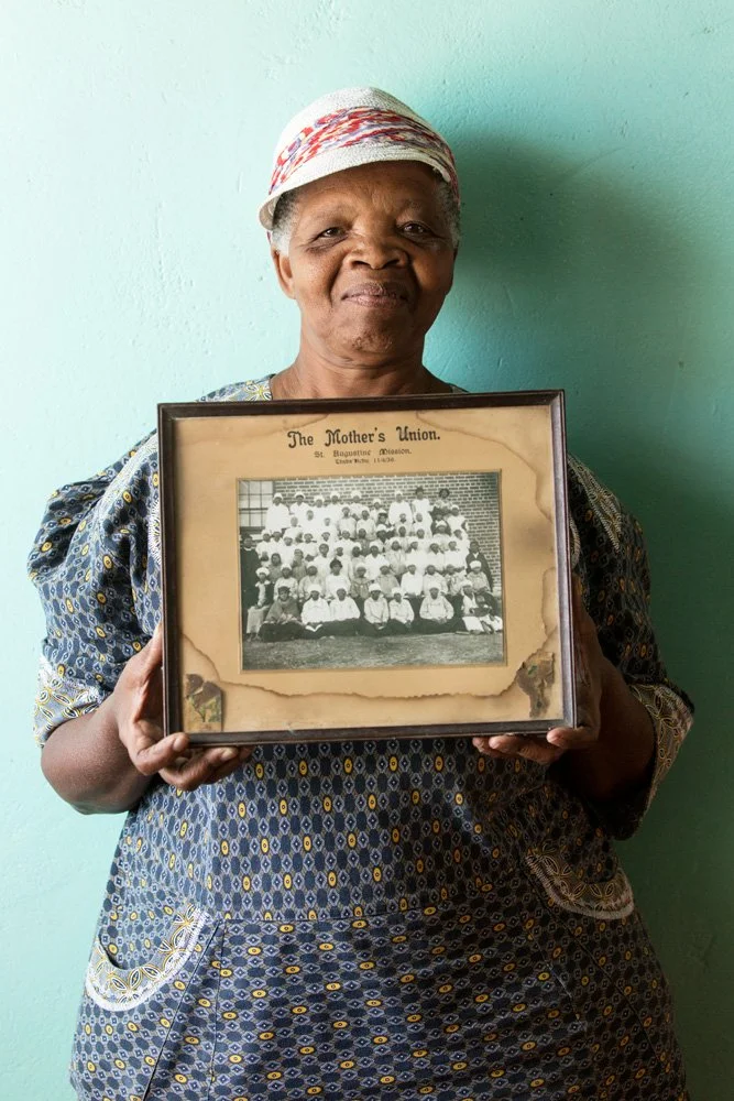 Dora Malefane holds a framed black-and-white photograph featuring her grandmother (1st row, 1st sitting from the left, with the black headscarf).  The group portrait depicts the ladies of The Mother's Union at the St. Augustine Mission in Thaba Nchu 