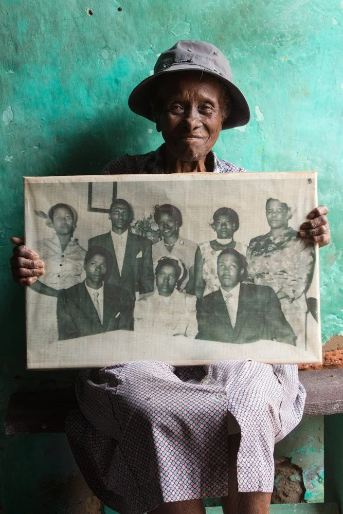 Bessie Moreki is a retired office clerk. She holds a plastic-wrapped digital enlargement of a black-and-white photograph, taken on her brother Joel Matheza 's wedding day in the mid-fifties. Bessie is the lady with the radiant smile, standing in the 