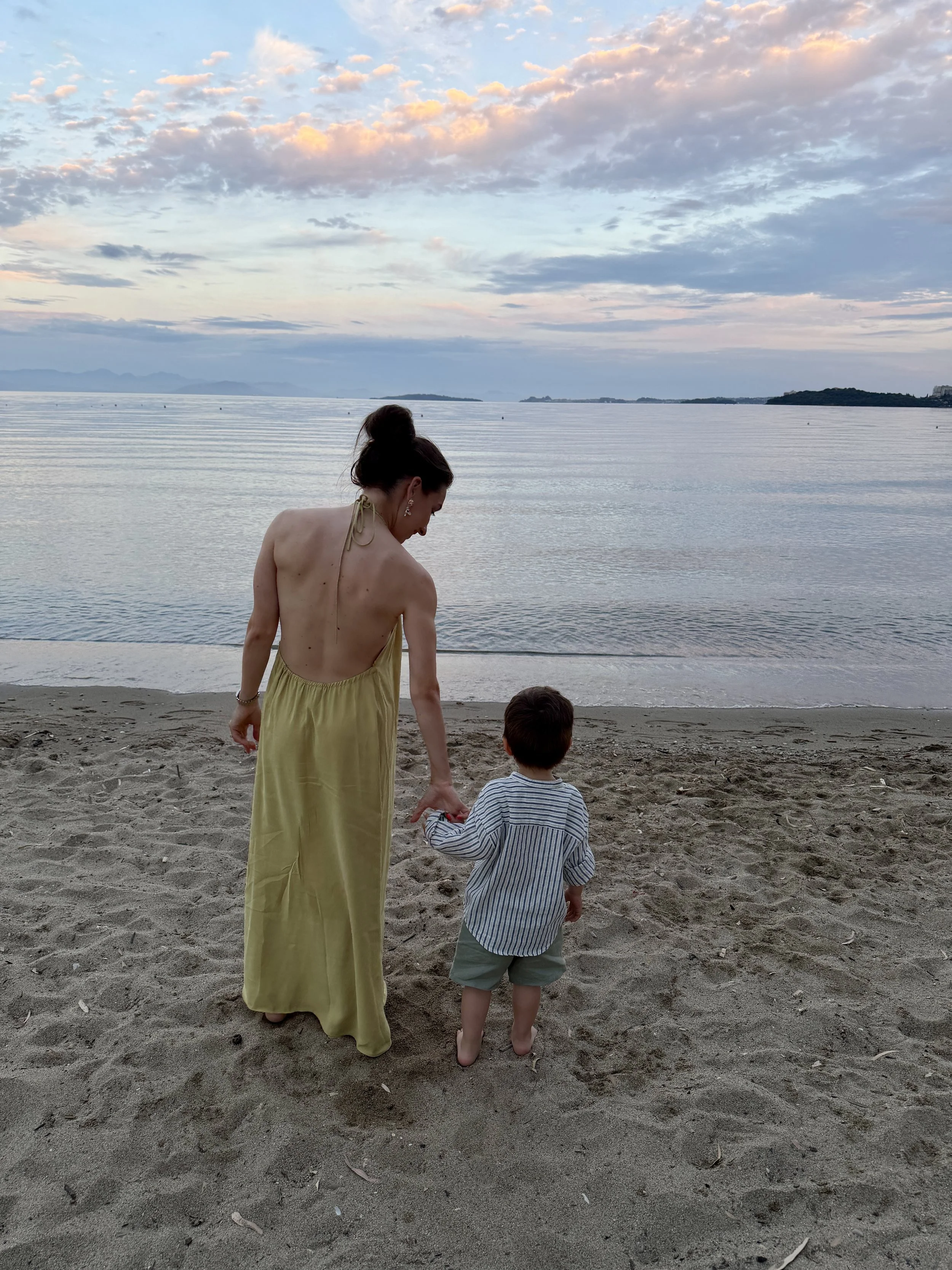 A woman in a yellow dress and a young boy in striped shirt and green shorts walking on a sandy beach by a calm body of water during sunset, with clouds and distant landforms in the background.