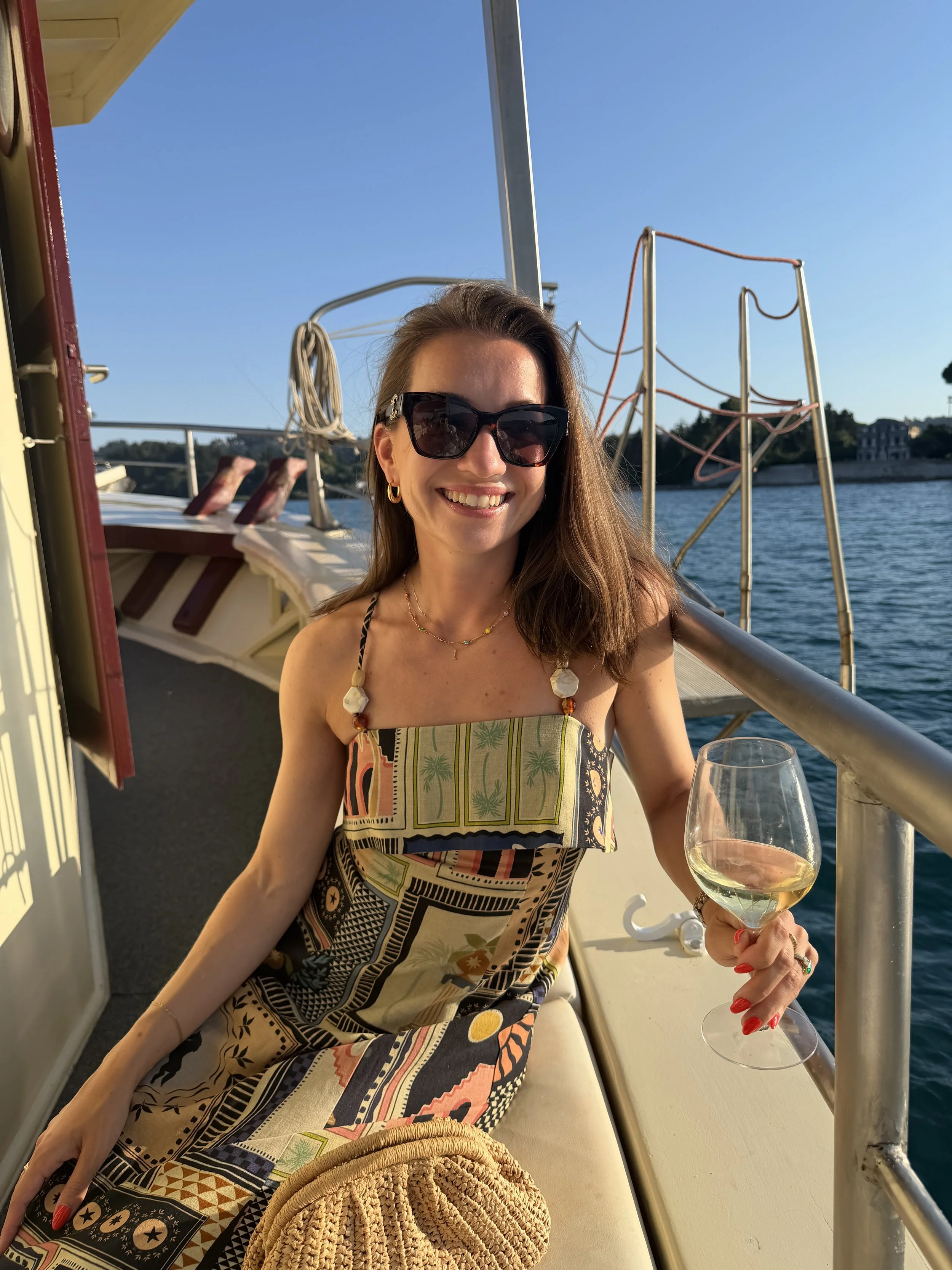 Woman with sunglasses holding a glass of white wine on boat with water and blue sky in background.