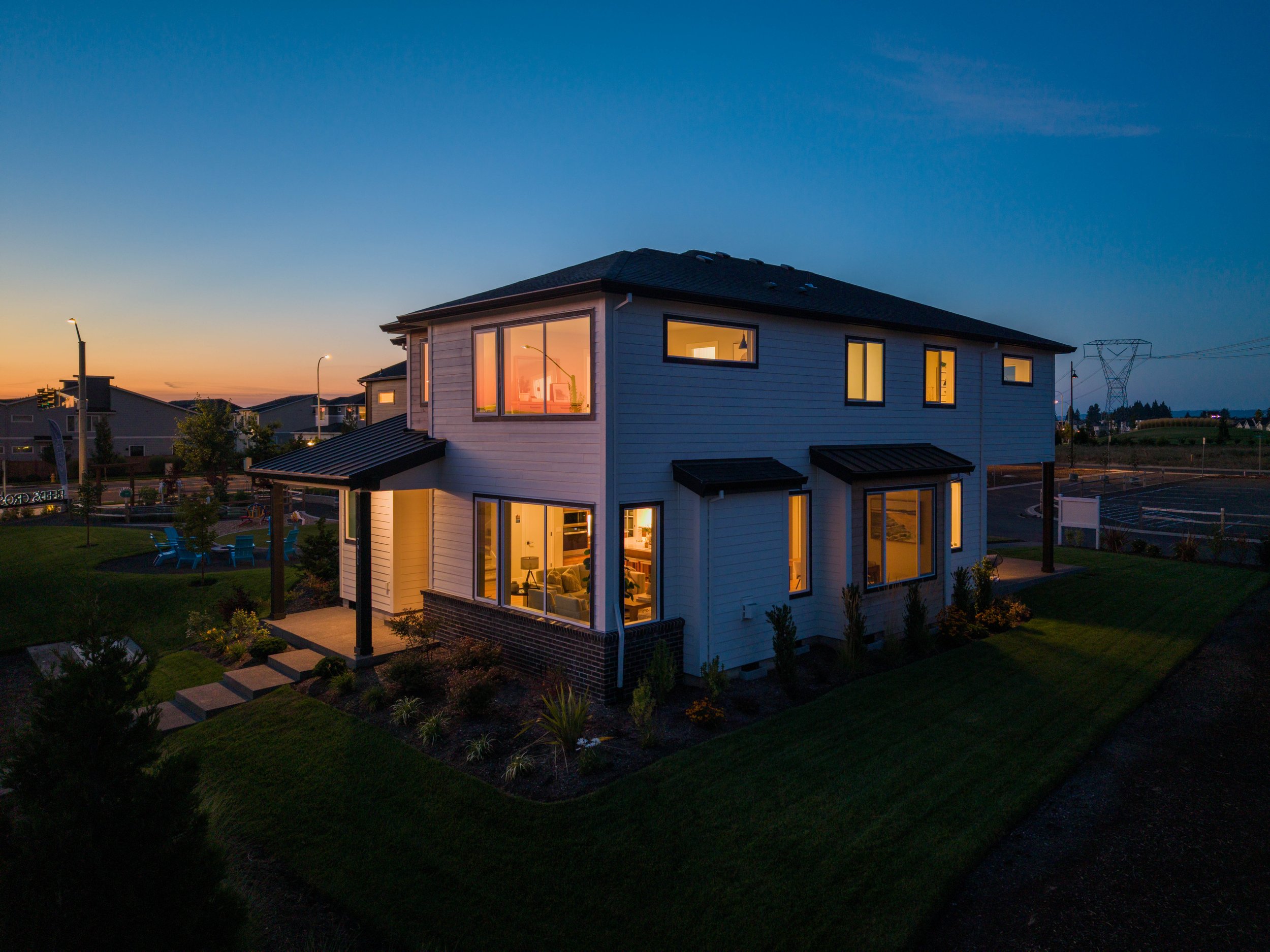 A modern two-story house illuminated from the inside, with large windows and a backyard with chairs, set against a dusk sky with a gradient of blue and orange.