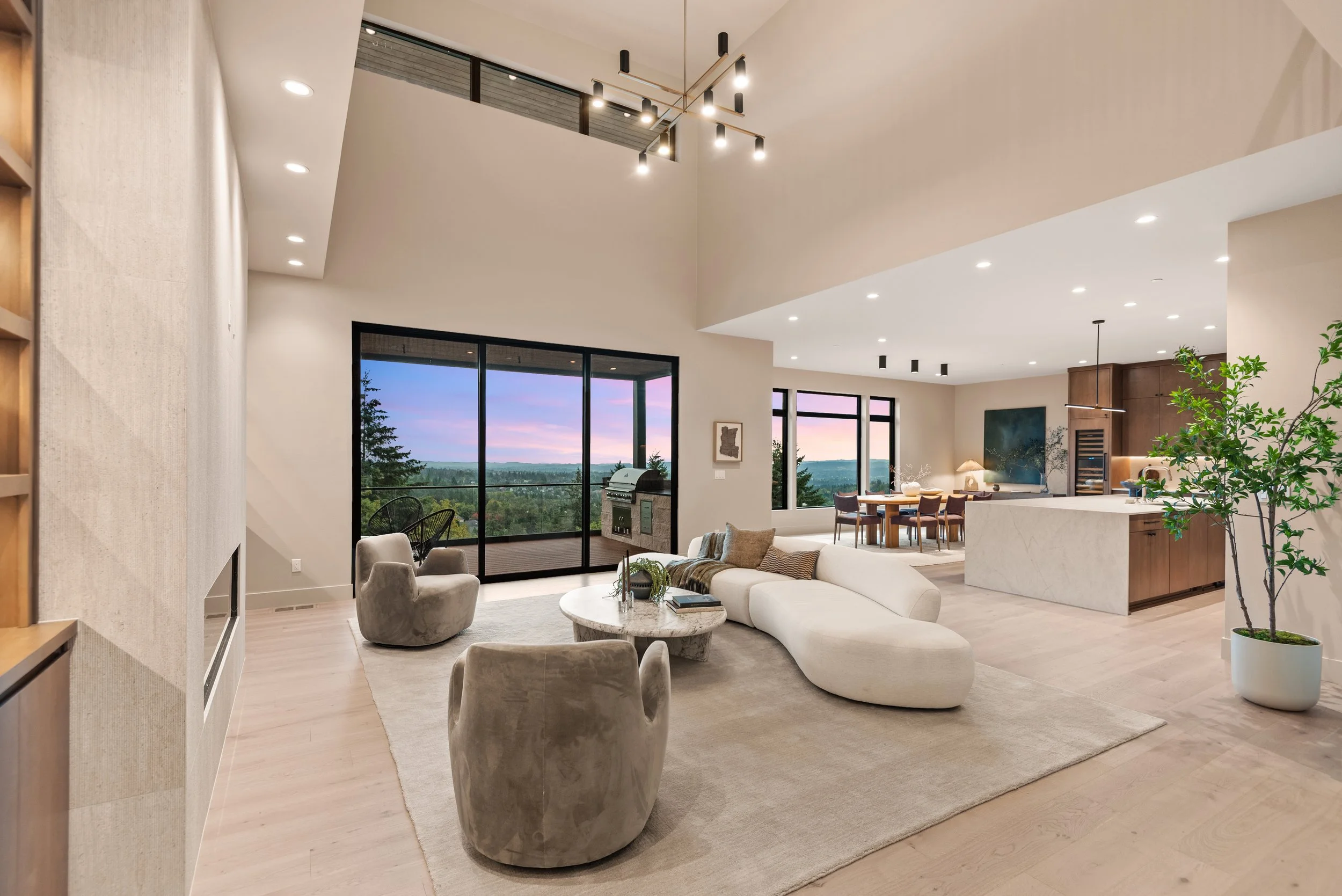 Modern open-concept living room and kitchen with large windows showing a scenic sunset view, featuring a white sofa, beige armchairs, a round coffee table, a dining table, and contemporary lighting fixtures.