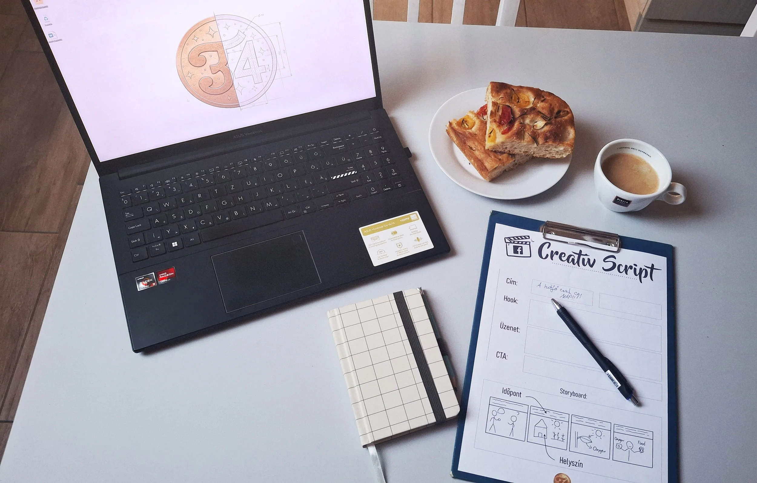Egy fehér asztalon laptop, egy szelet pizzát tartalmazó tányér, egy csésze kávé, egy sztoribord-munkalappal ellátott írótábla, egy kis jegyzetfüzet és egy toll helyezkedik el.