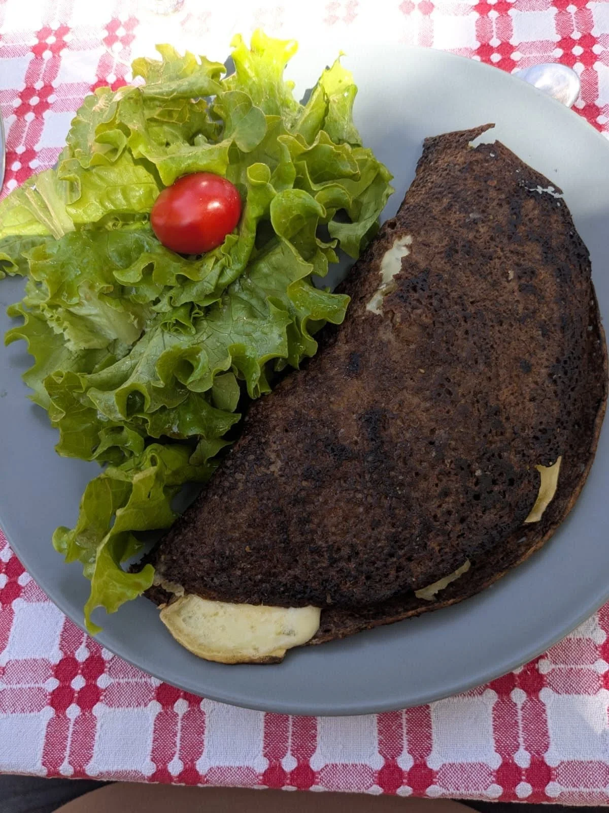 Une assiette avec une crêpe noire au chocolat et une salade verte avec une tomate cerise, posée sur une nappe rouge à carreaux blancs.