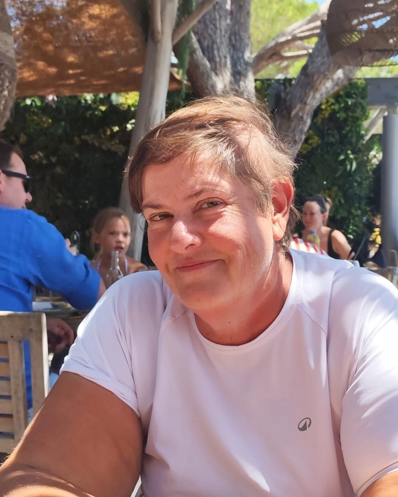 Un homme souriant avec des cheveux châtains, porté un t-shirt blanc, assis à une table en extérieur pendant la journée, avec des gens en arrière-plan et des arbres.