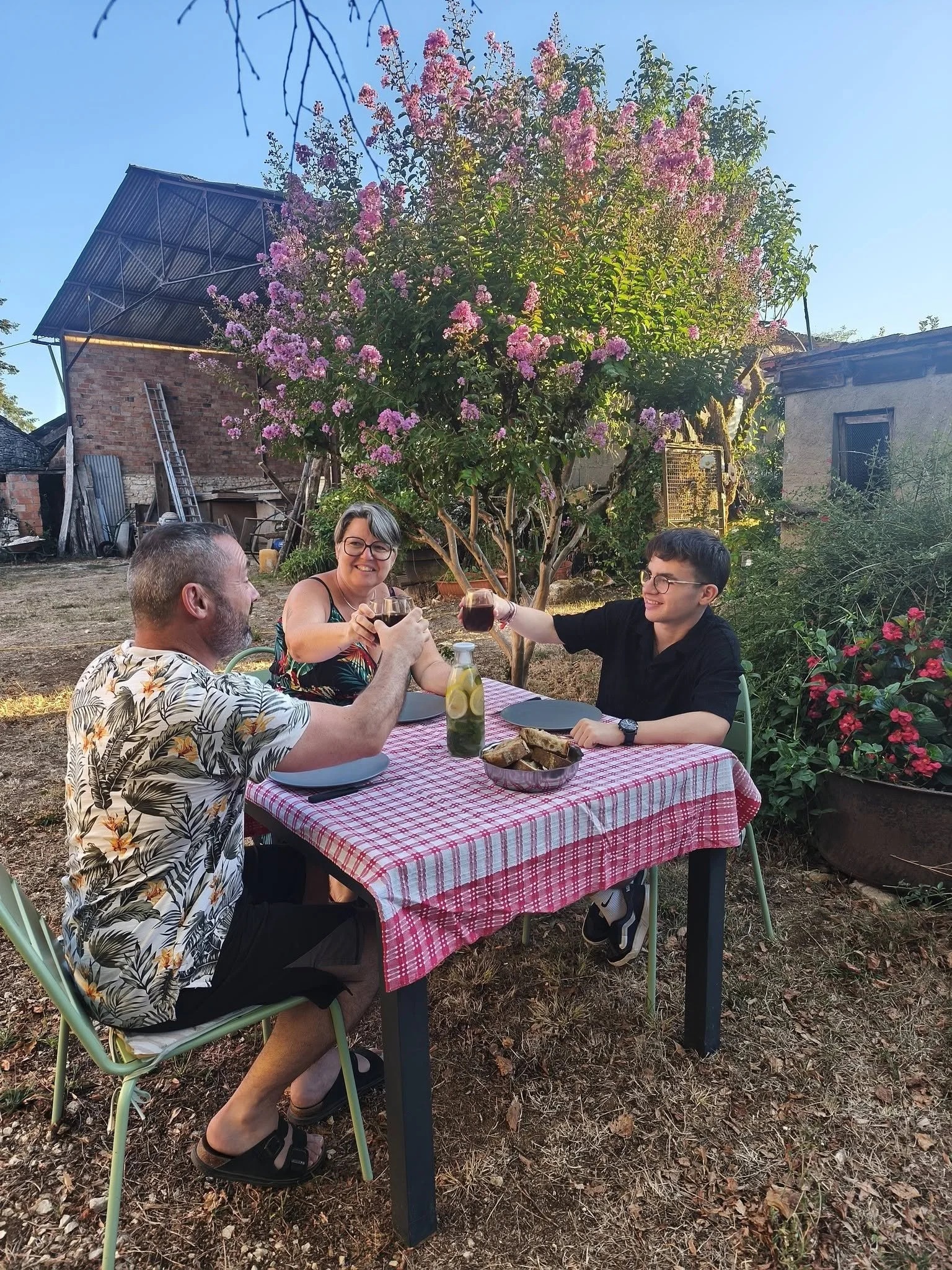 Trois personnes, deux hommes et une femme, assises autour d'une table en extérieur, à l'ombre d'un arbre en fleurs roses, en train de boire un verre de vin. La table est recouverte d'une nappe à carreaux rouges et blancs, avec deux assiettes et une bouteille d'eau citronnée. Le fond montre une maison en pierre, un toit en tôle, et un espace verdoyant.