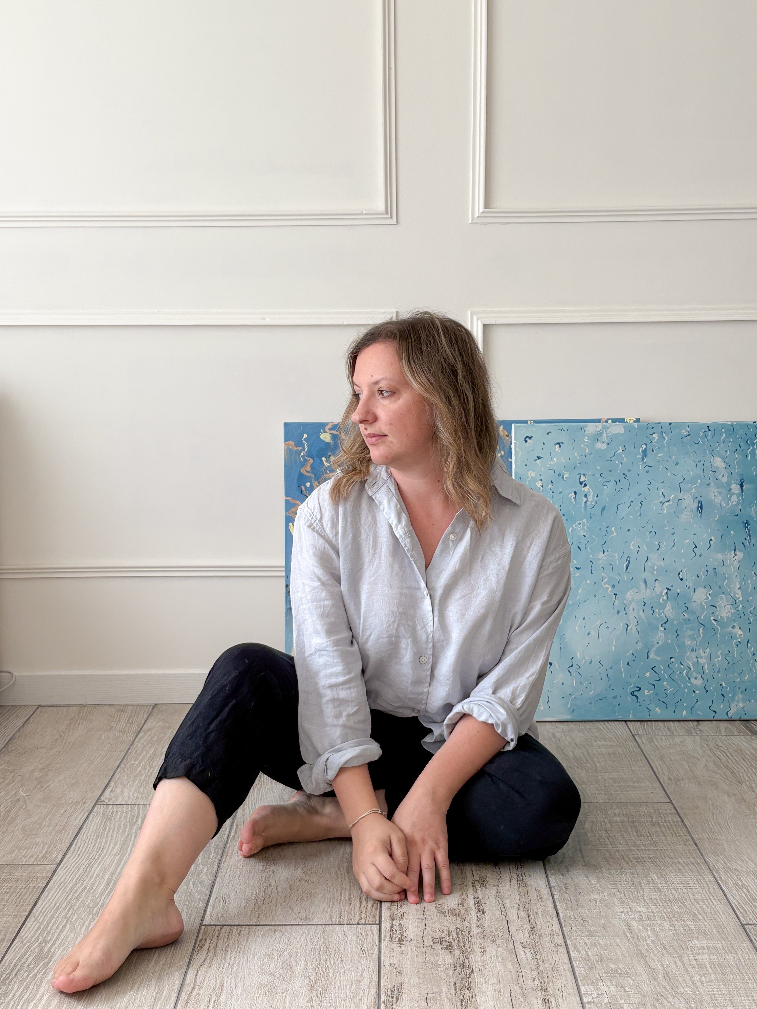 A woman artist with wavy brown hair and a light shirt is sitting on the floor with her legs crossed. Behind her, there are two abstract blue paintings leaning against a white wall with decorative molding.