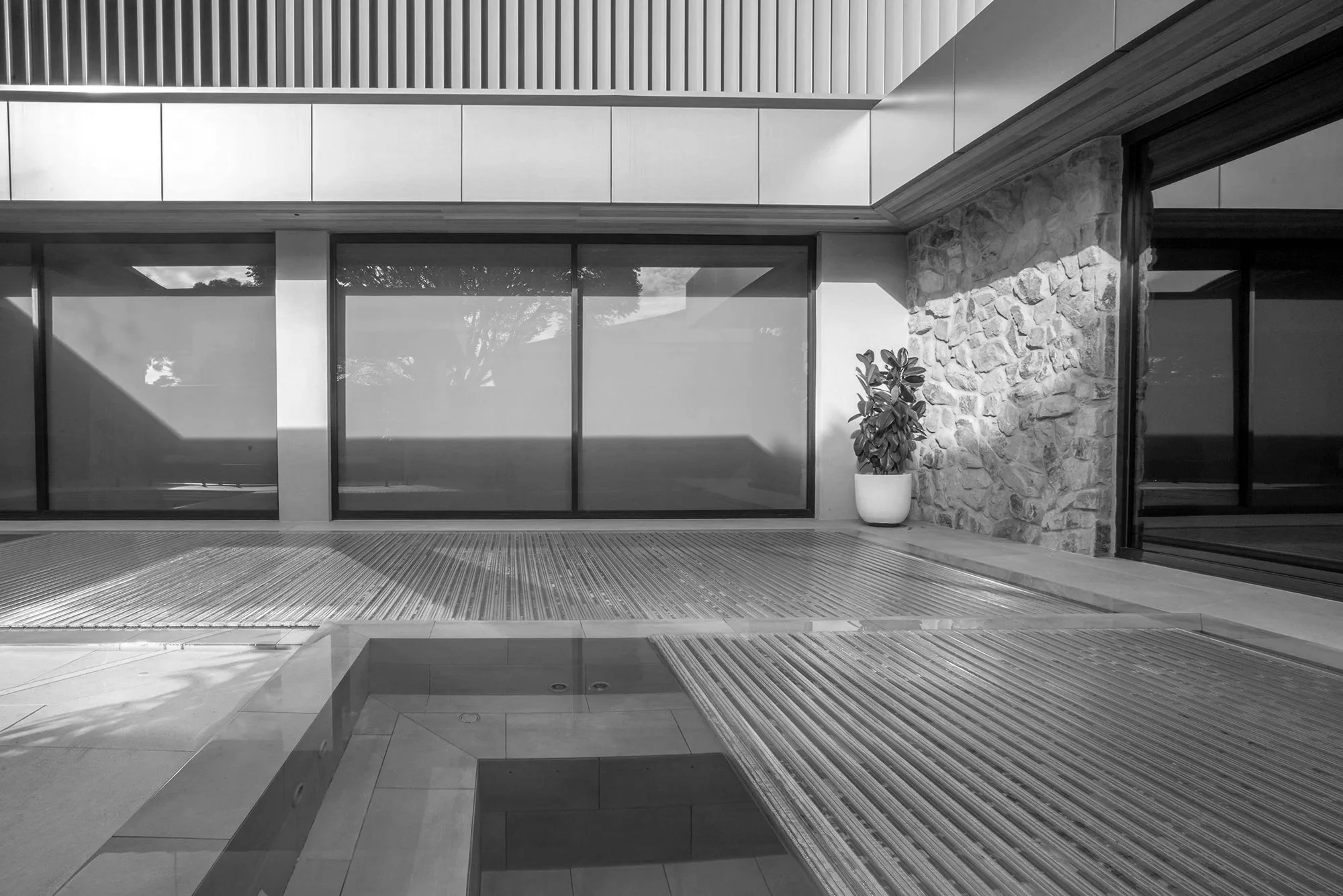 Modern courtyard with a shallow reflecting pool, striped decking, floor-to-ceiling windows, stone wall, and potted plant
