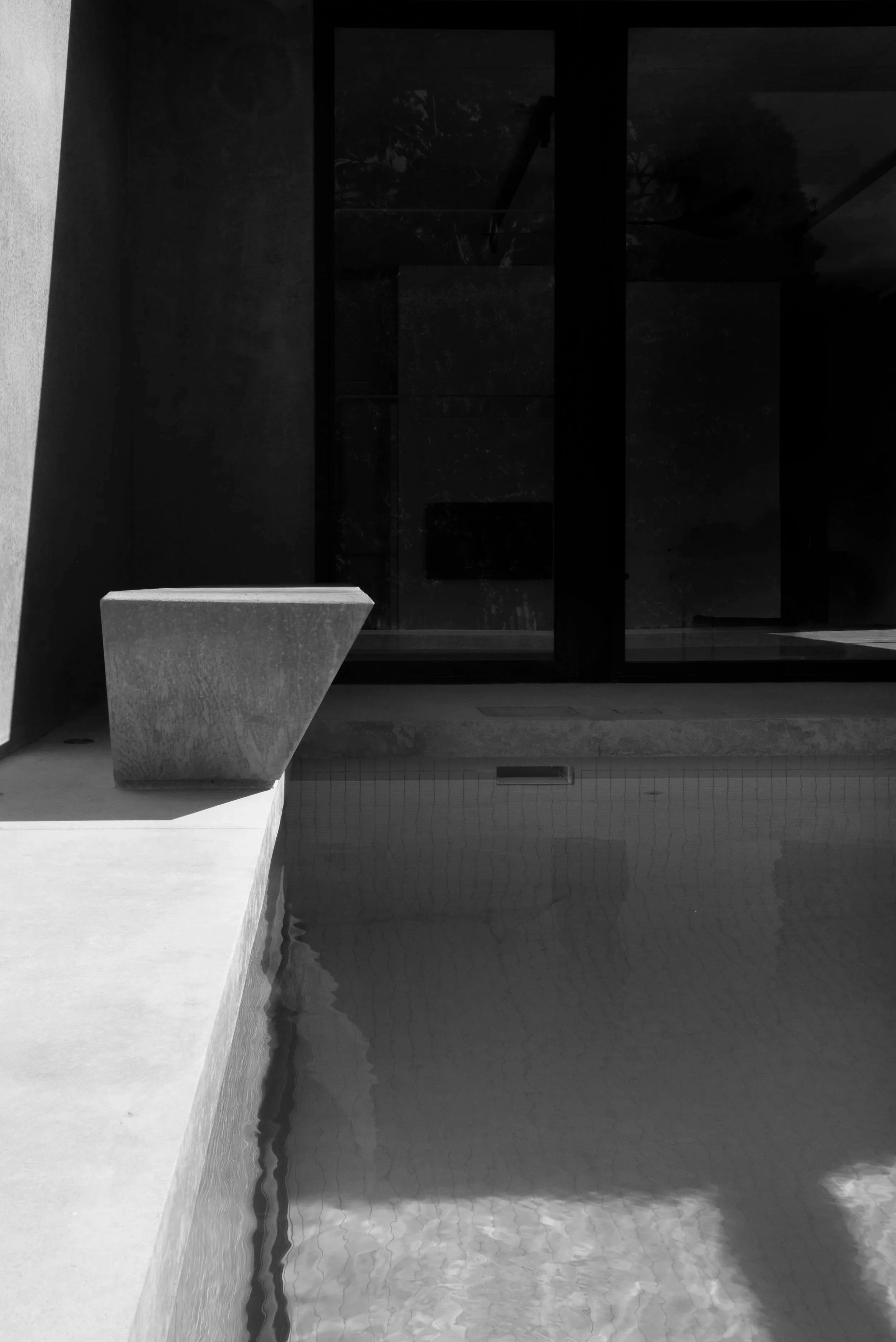 Dark modern indoor pool with a concrete bench, reflecting water, and large windows.