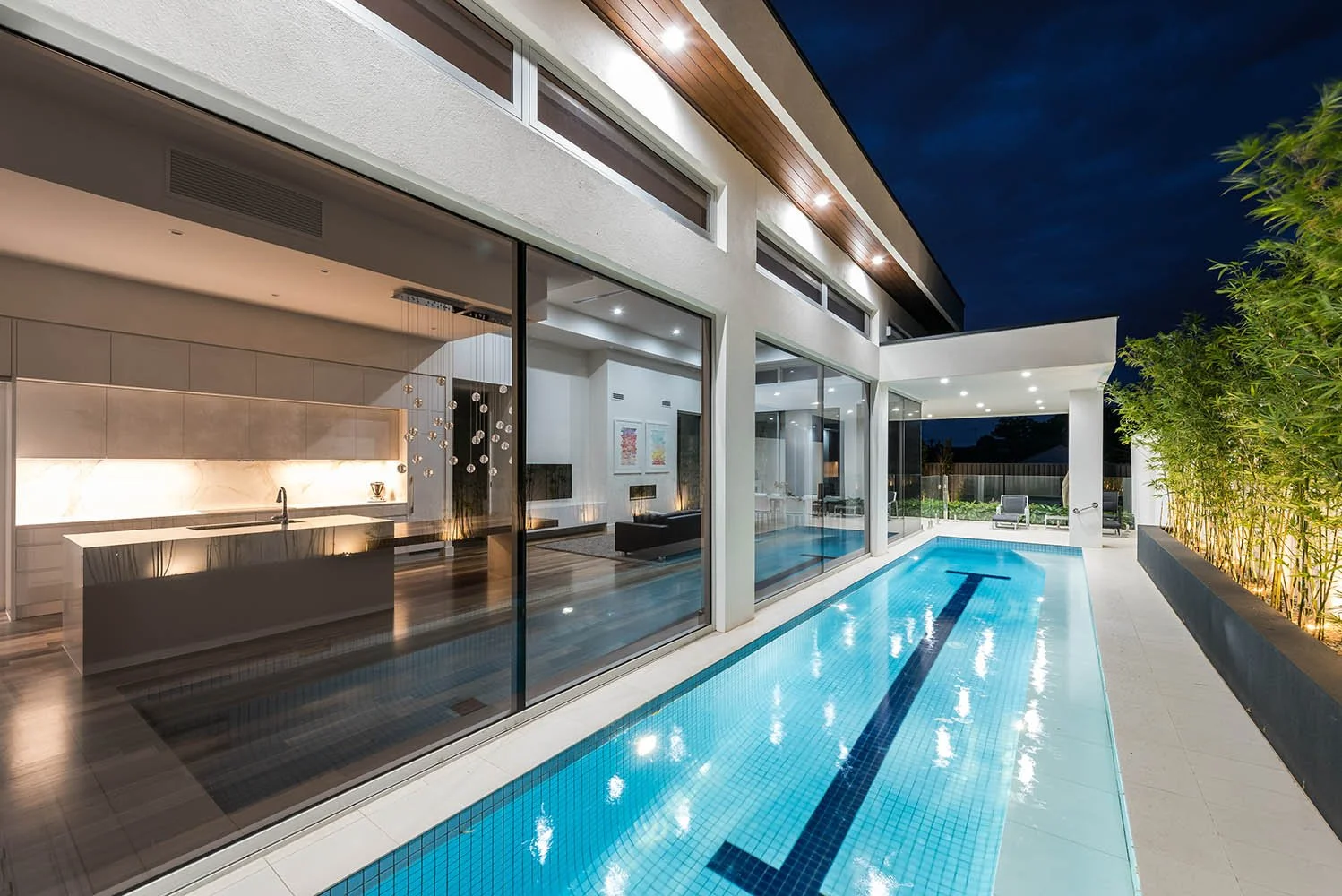 Modern luxury home with illuminated indoor-outdoor pool, open-plan kitchen, and floor-to-ceiling glass walls at night
