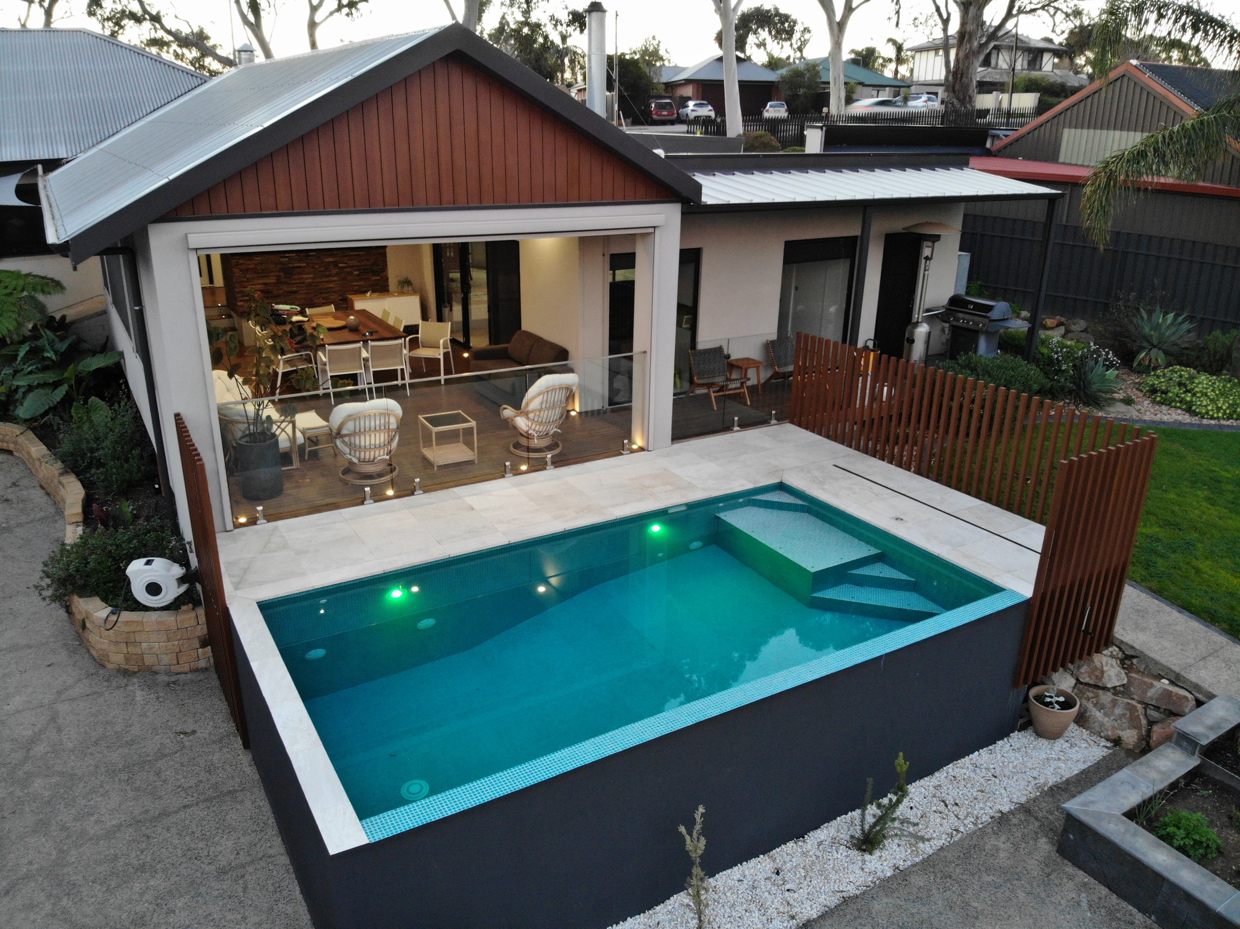 Modern backyard with a lit swimming pool, covered outdoor dining area, patio seating, barbecue grill, and landscaped garden