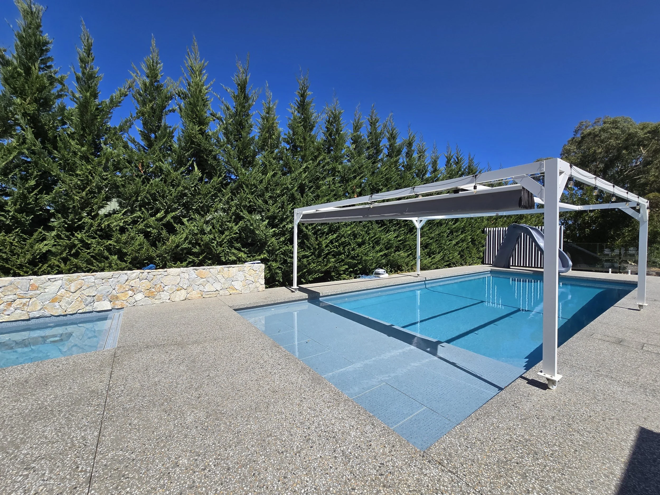 Outdoor swimming pool with a shaded pergola, water slide, spa, stone retaining wall, and tall evergreen trees under a clear blue sky