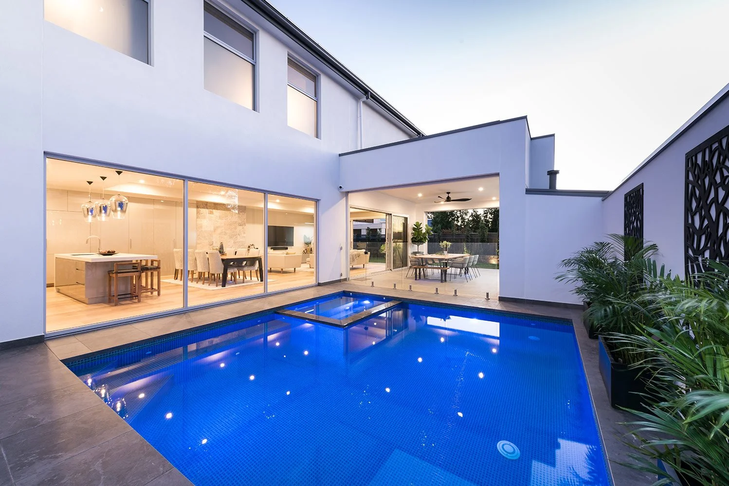 Modern luxury home with a blue-lit courtyard swimming pool, glass walls, open kitchen, dining area, and covered outdoor patio
