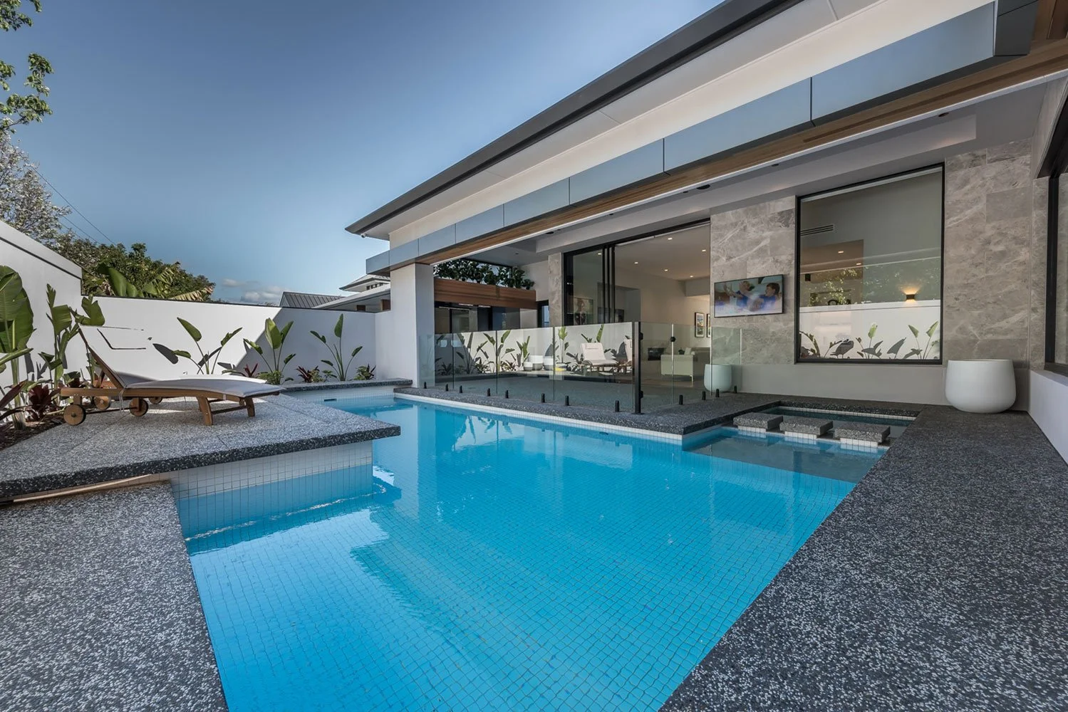 Modern luxury home with a turquoise swimming pool, spa, glass walls, lounge chairs, and tropical landscaping