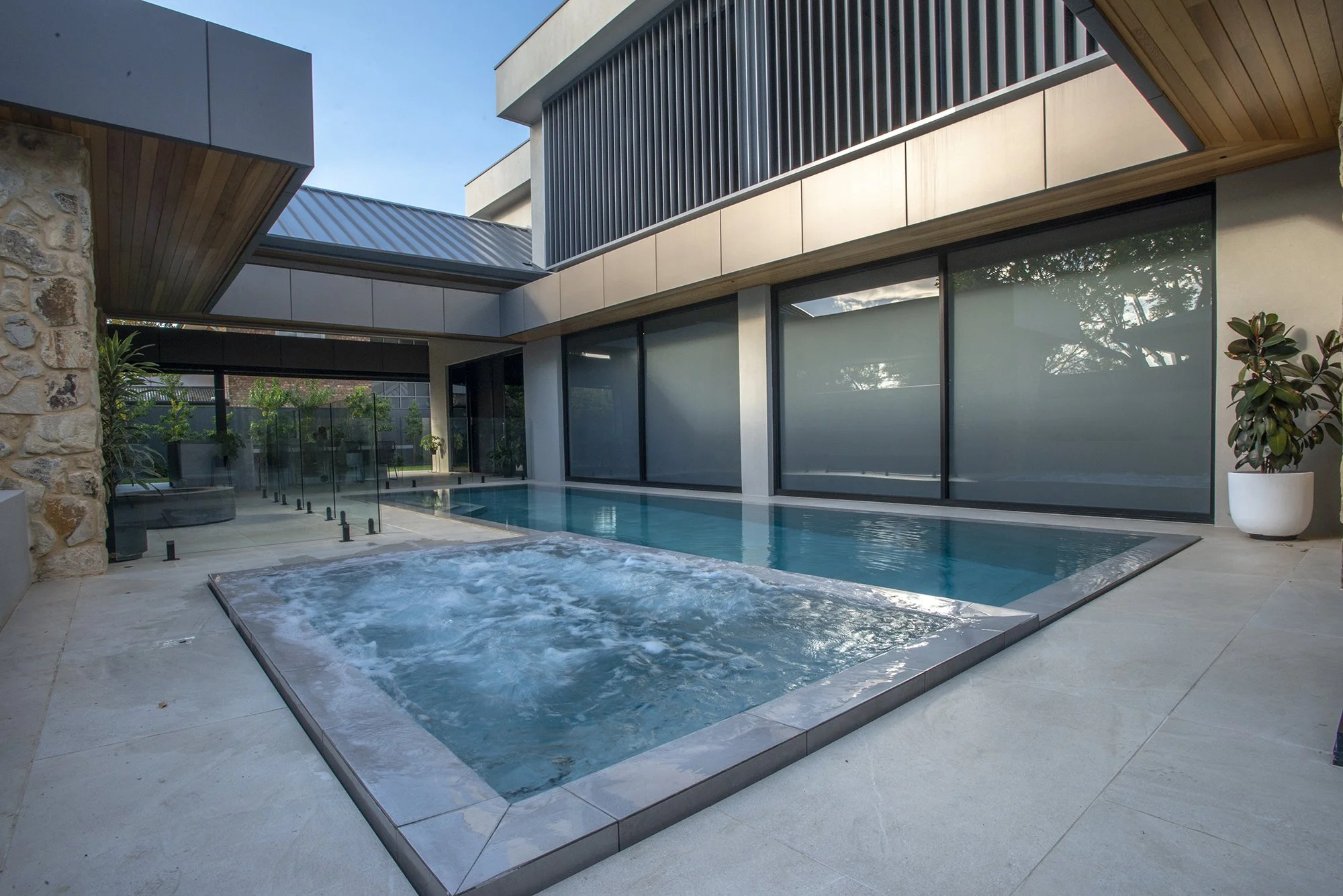 Modern home courtyard with a swimming pool and bubbling spa, floor-to-ceiling windows, stone walls, and potted plants