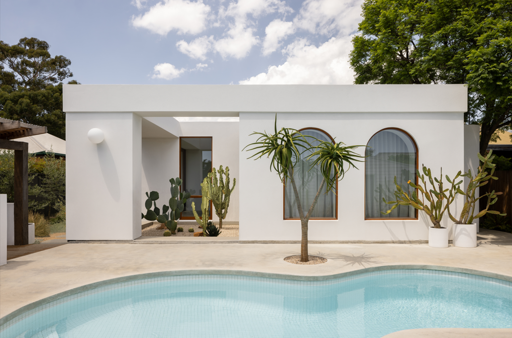 Modern white stucco home with arched windows, potted cacti, tropical plants, and a curved swimming pool